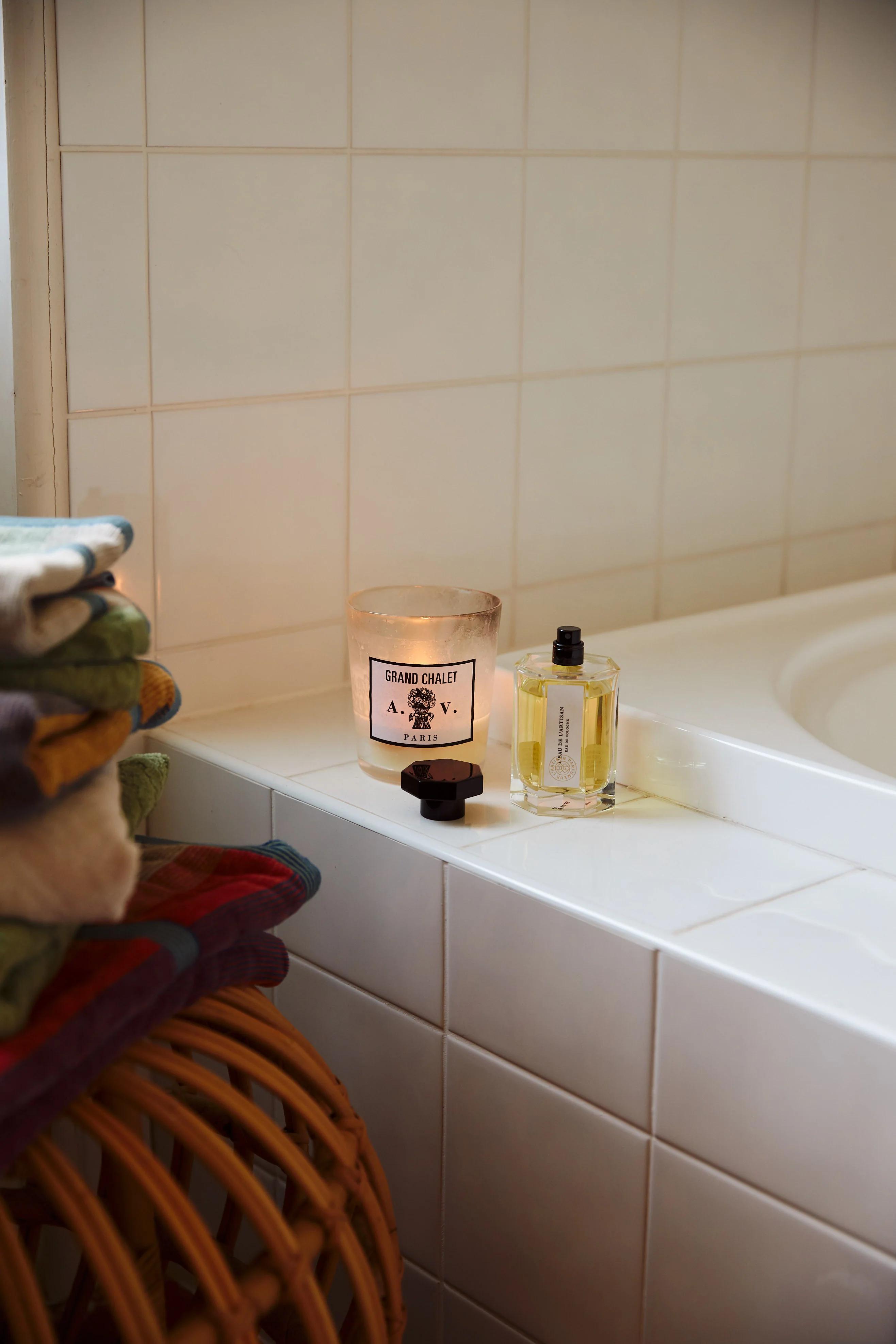 Lit candle and yellow liquid bottle on white tiled bathtub edge. Wicker basket with folded colourful towels nearby.