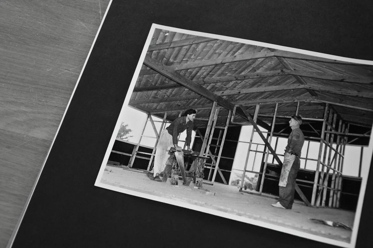 Black and white photo: Two men constructing wooden building. One at saw, other observing. Exposed beams and scaffolding visible. Photo on black mat.