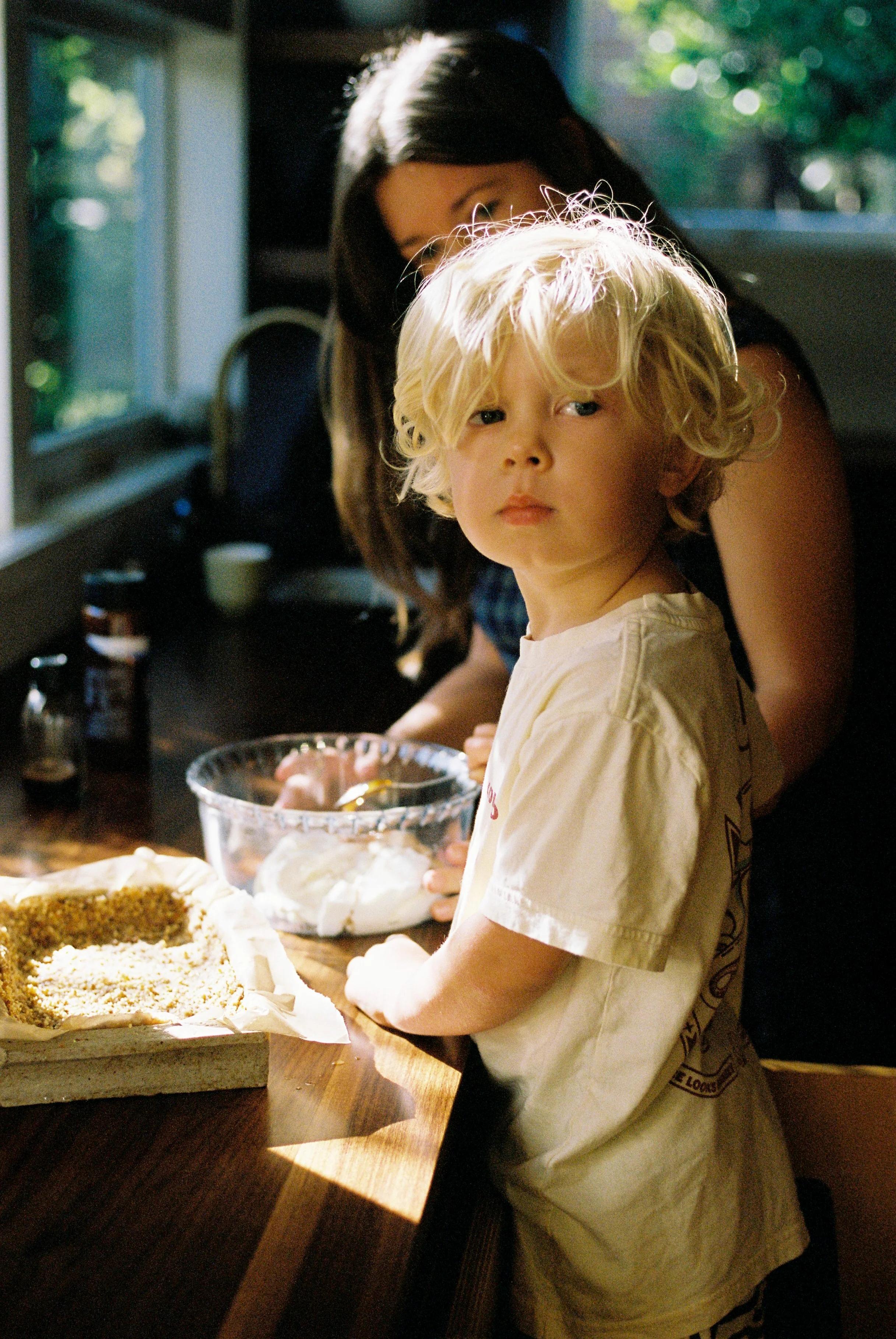 Blonde child at counter with woman holding bowl. Natural light through window. Midcentury home atmosphere.