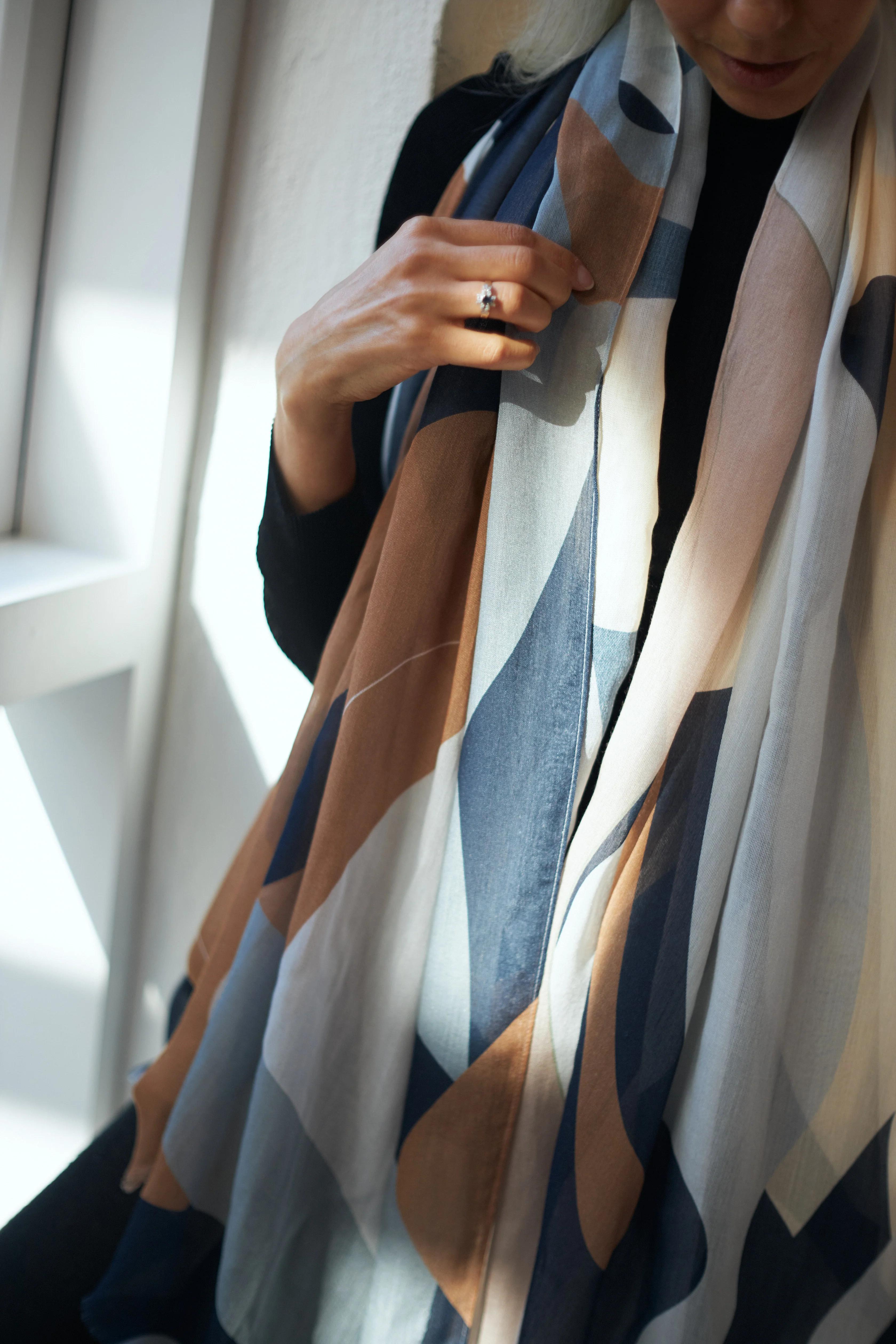 A person wearing a dark-coloured outfit draped in a large, abstract-patterned scarf with shades of brown, blue, and cream. They hold the scarf with one hand near their chest, showing a ring on their finger. Soft, natural light filters through a window beside them.