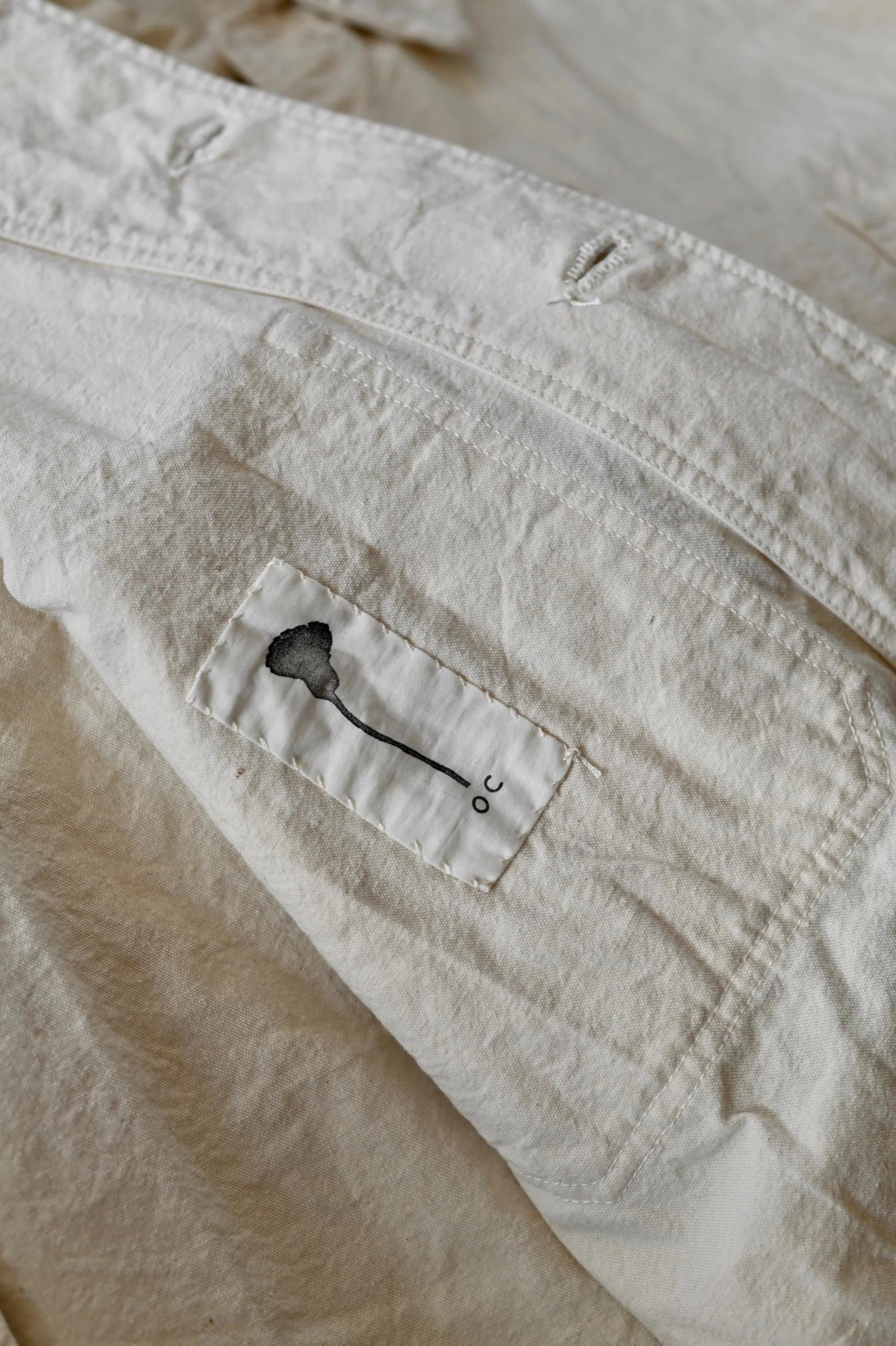 Close-up of light fabric with stitched patch. Black embroidered key-like figure and "c". Likely from Oliver Church's Singular Garments.