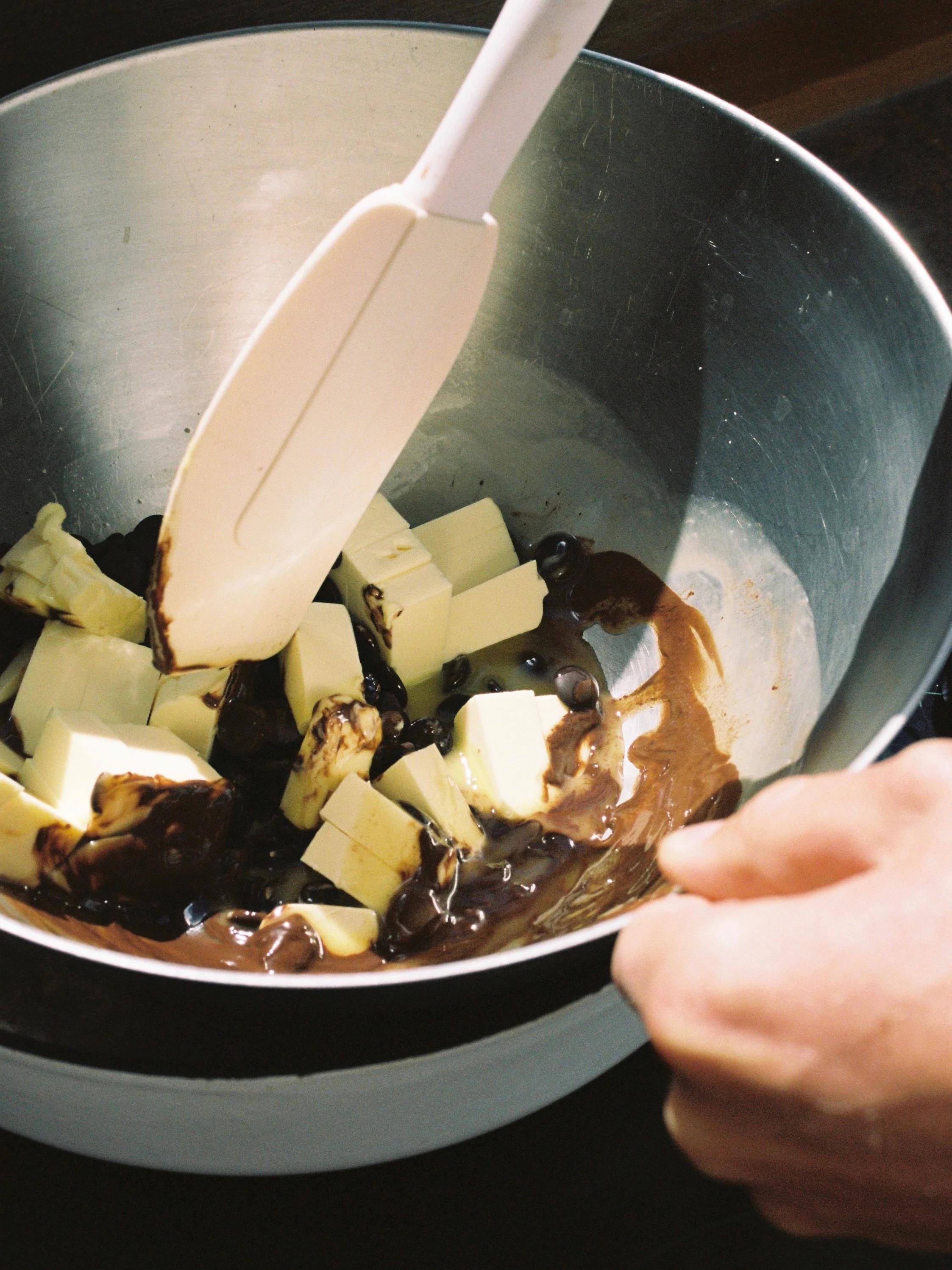 Close-up of someone stirring melted chocolate and butter in a metal bowl using a white spatula, using the double boiler method for a festive cake.