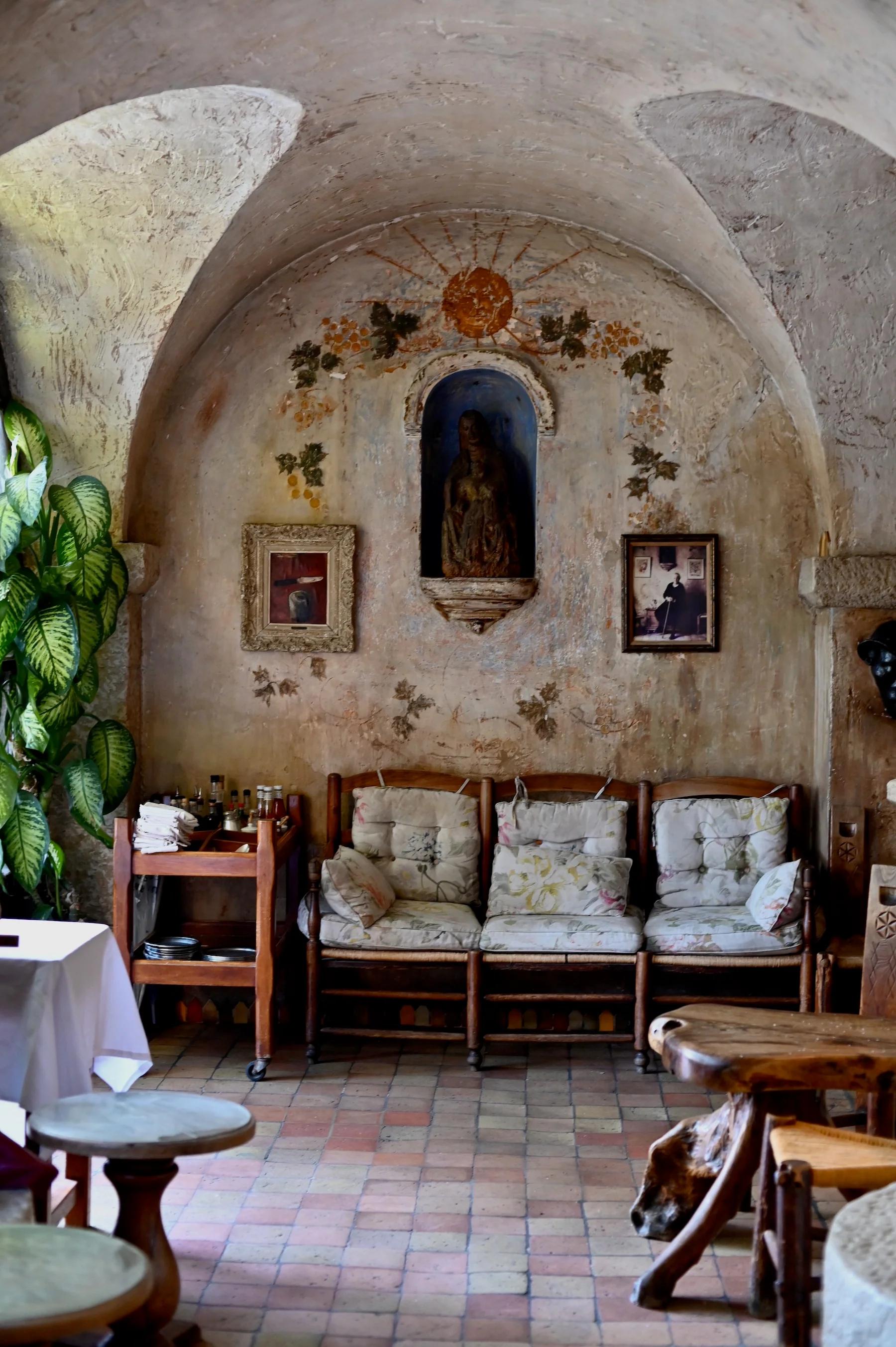 Rustic seating area with arched ceilings and stone alcove housing a small statue. Framed pictures and ivy adorn walls, with wooden chairs nearby.