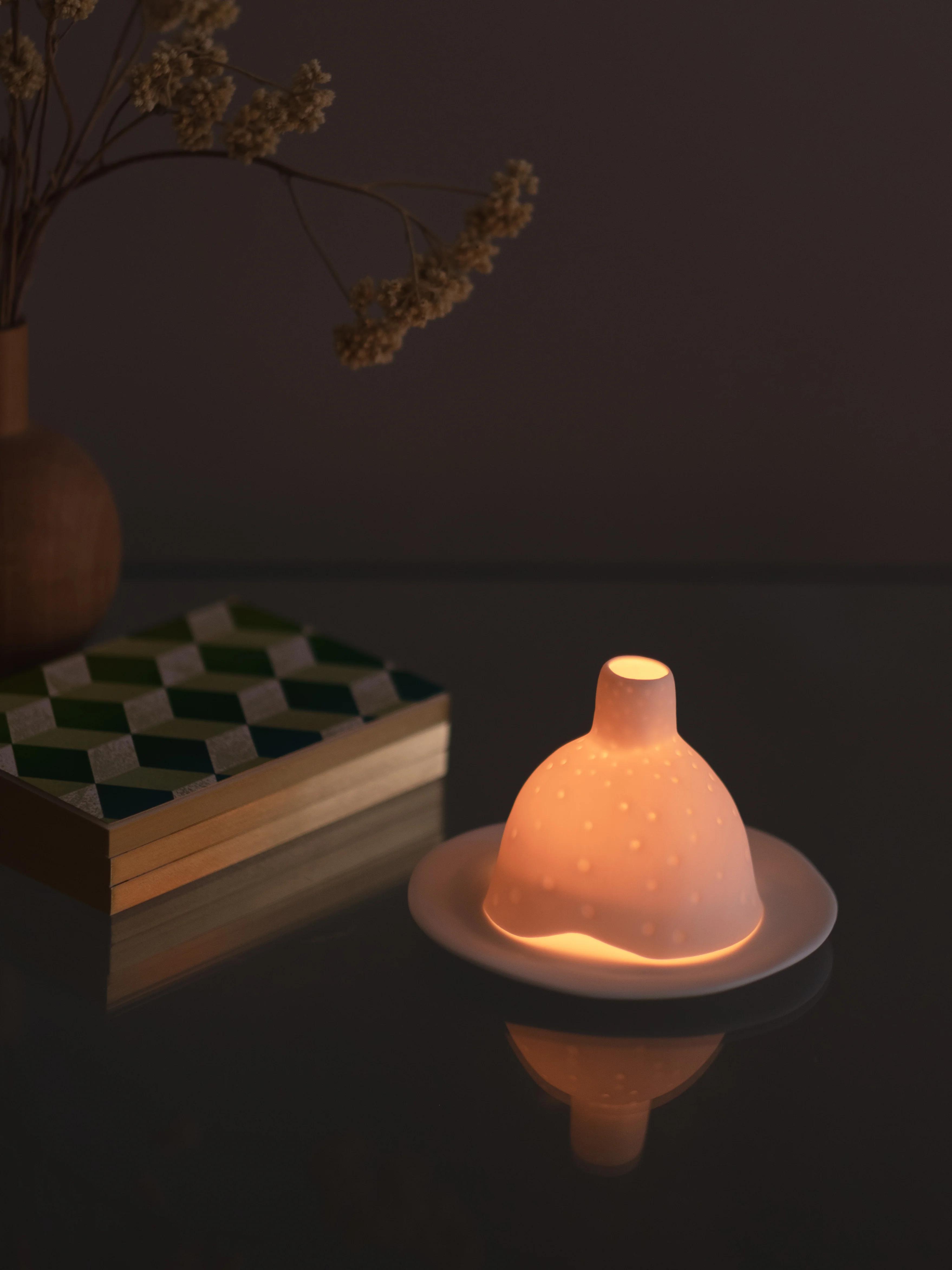 Soft, dimly-lit tulip-shaped lamp with dotted pattern on a glass surface. Nearby, a geometric-patterned book and wooden vase with dried flowers create a cozy ambiance.