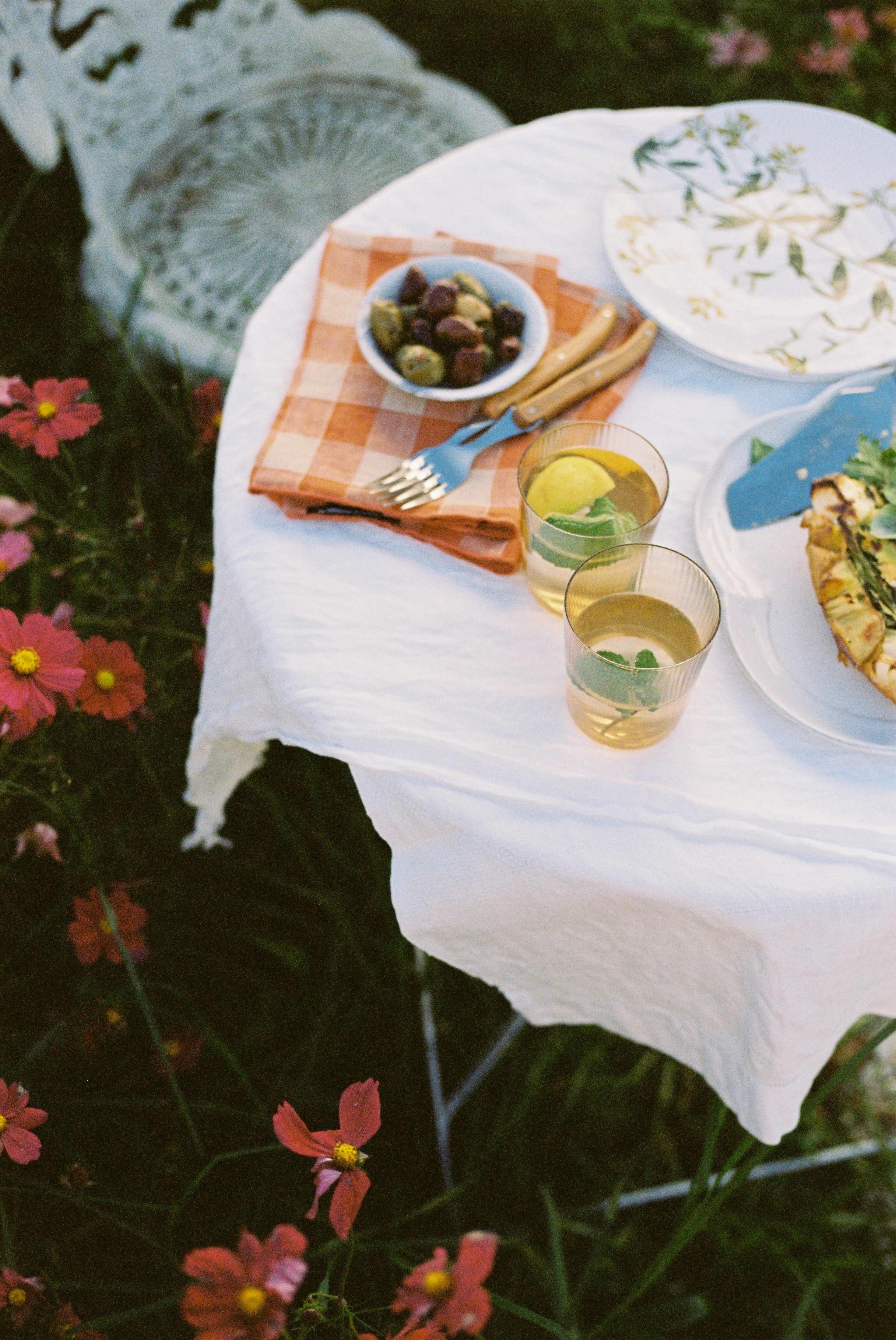 Garden table with white cloth, plaid napkin, cutlery, fruit bowl, salad plate, and lemon water. Pink flowers bloom nearby, perfect for a spring celebration.