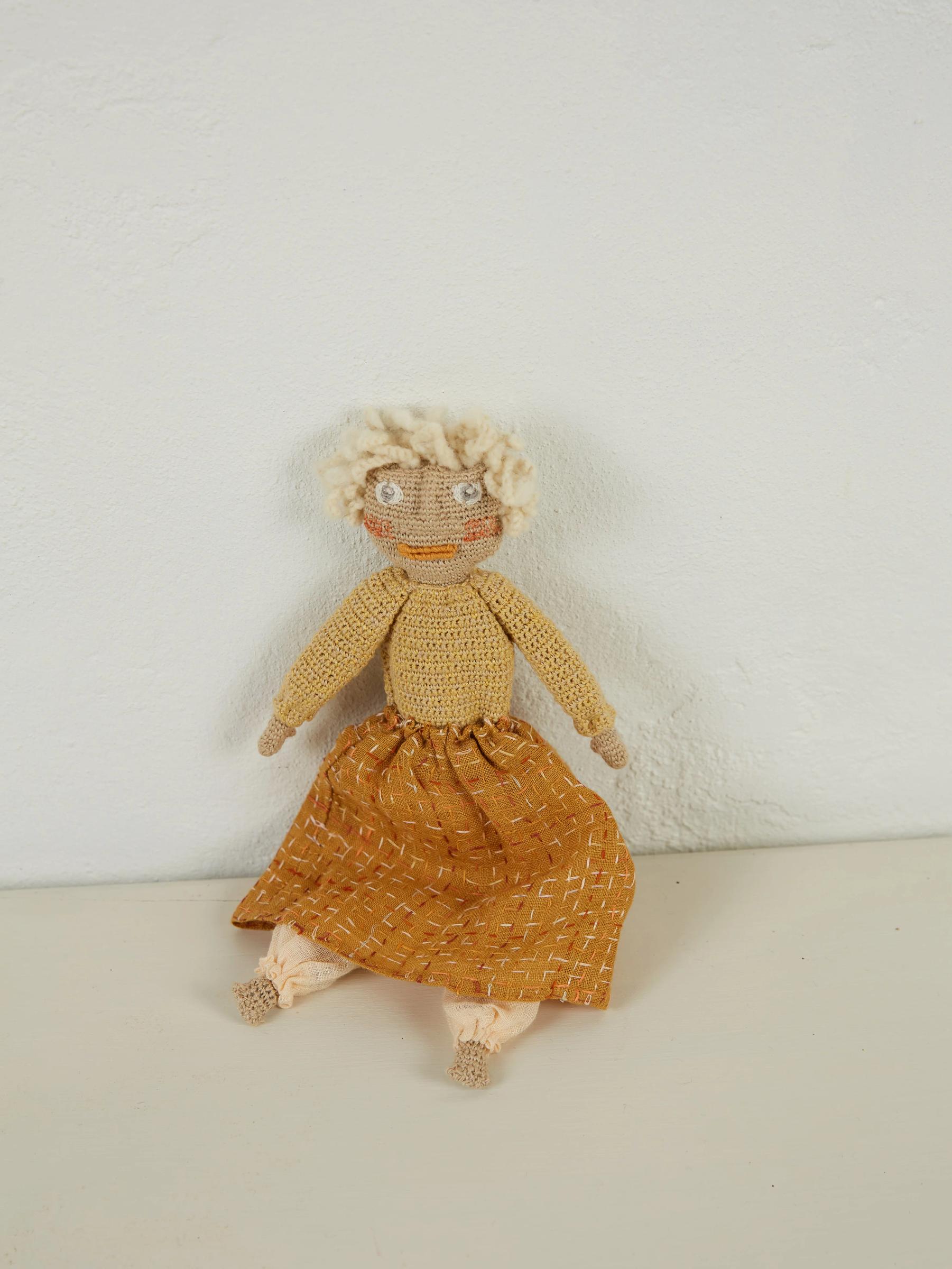 Handmade knitted doll with curly white hair in beige top and orange patterned skirt. Seated against light wall, legs extended. Reminiscent of Sophie Digard's style.