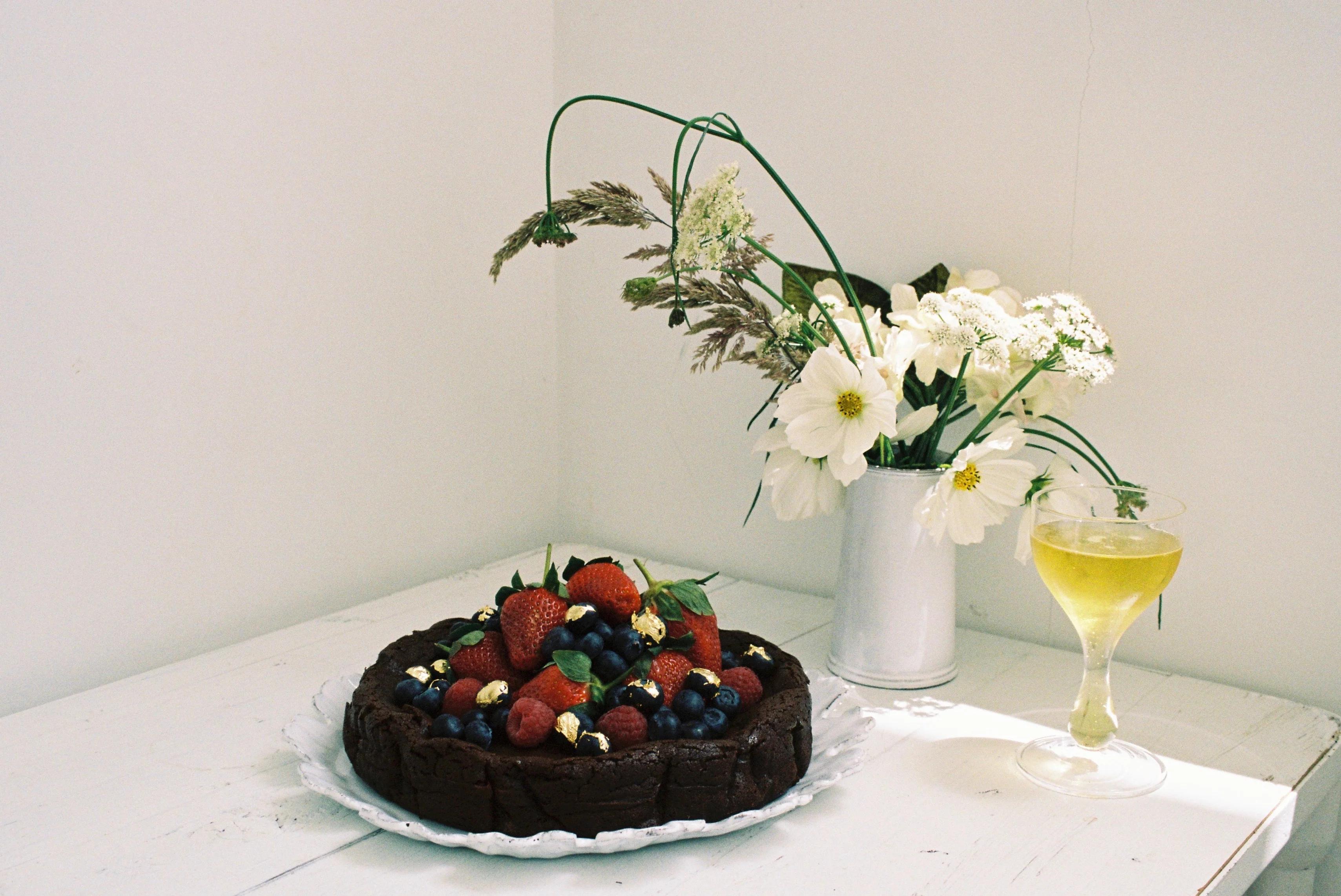 A flourless chocolate cake topped with strawberries, blueberries, and nuts on a white plate, with a glass of wine and a vase of white flowers in the background.