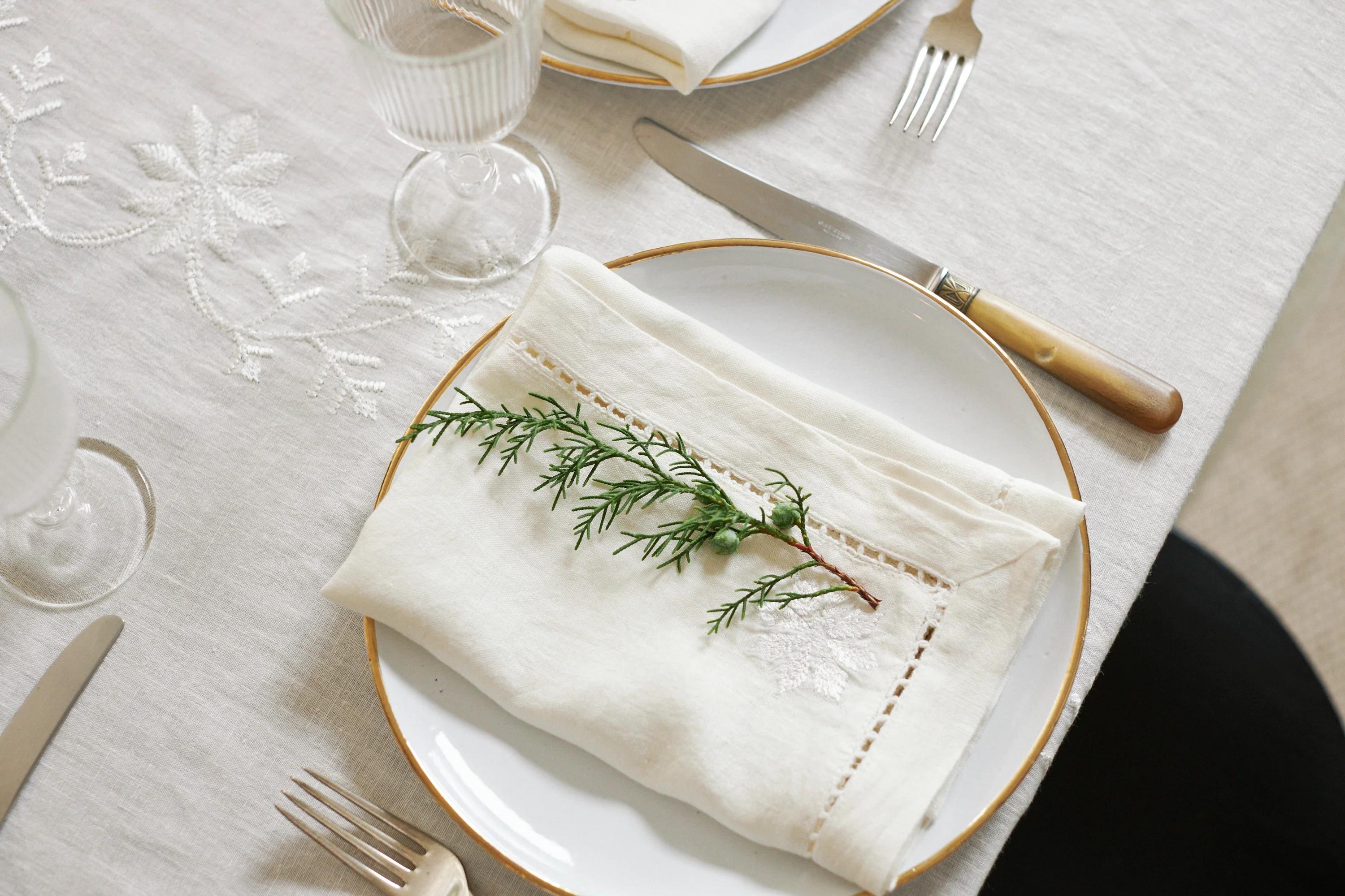 An aerial view of a place setting with a white plate, folded napkin, and greenery. Cutlery and glasses sit on an embroidered tablecloth.