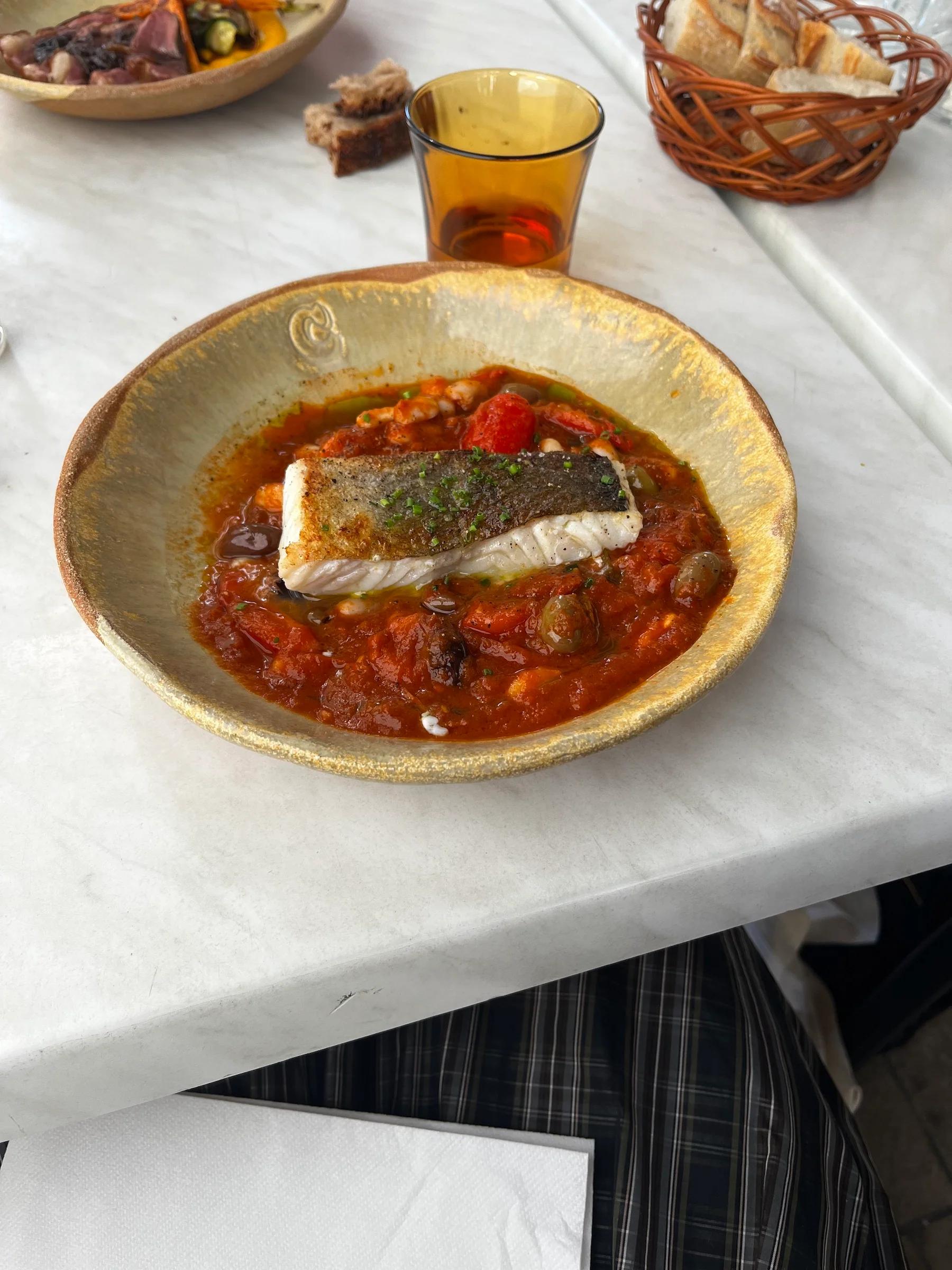 Ceramic bowl with grilled fish on tomato sauce and vegetables. Bread basket and amber glass visible, reminiscent of Marseille flavours.