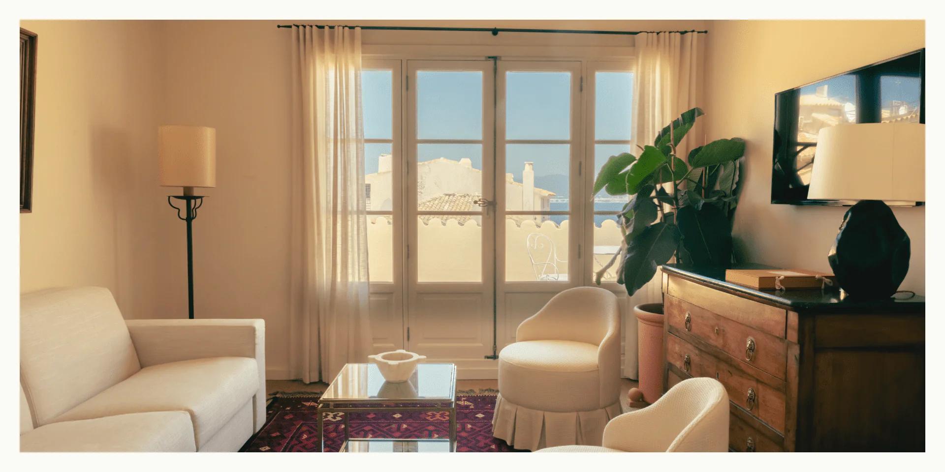 Living room with white sofa, chairs, wooden chest. Glass coffee table on colorful rug. Sheer curtains, Balthus Lamp, potted plant.