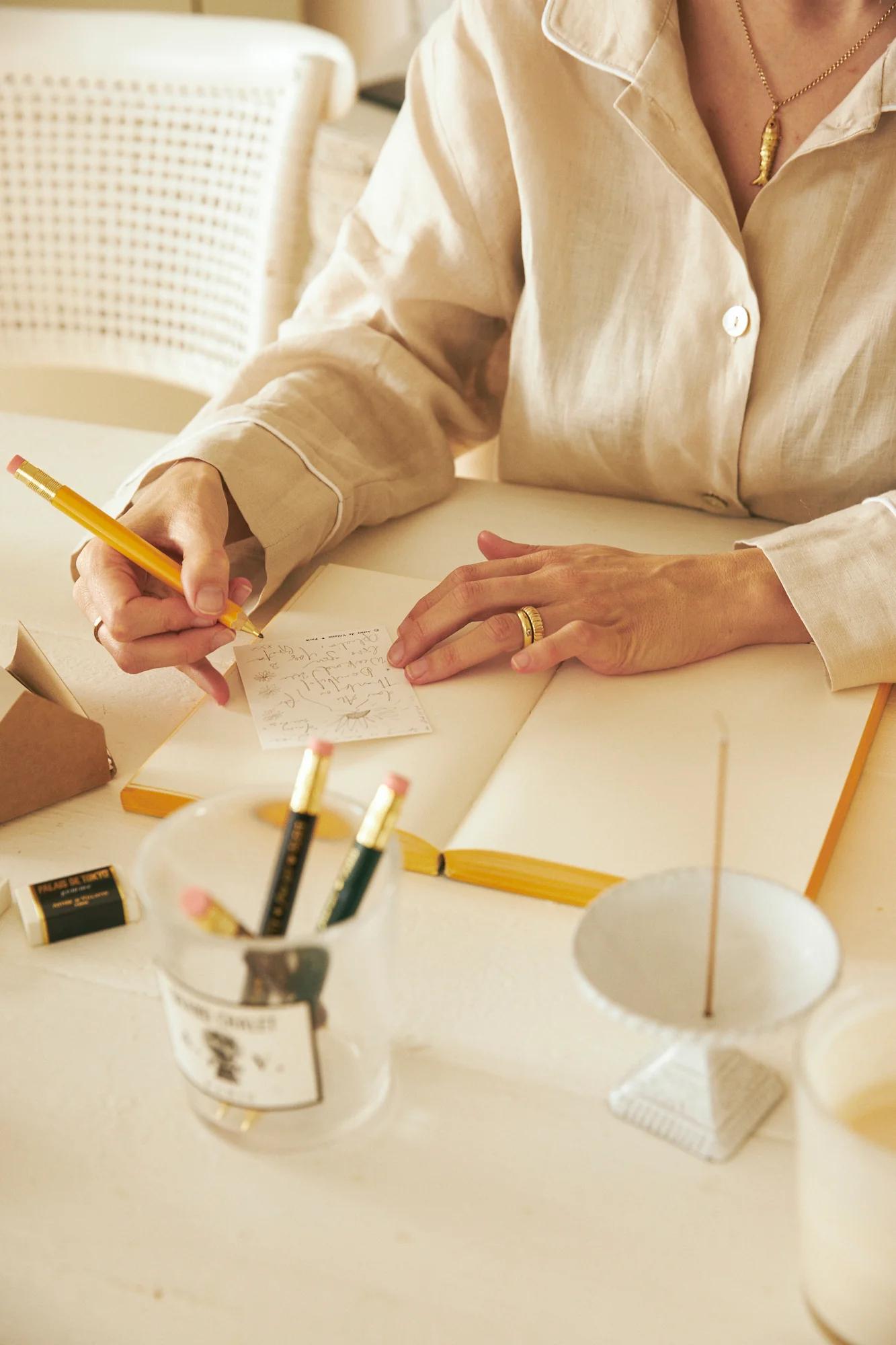 Person in light shirt writing cards with yellow pencil. Other pencils, eraser, and candle holder on table.