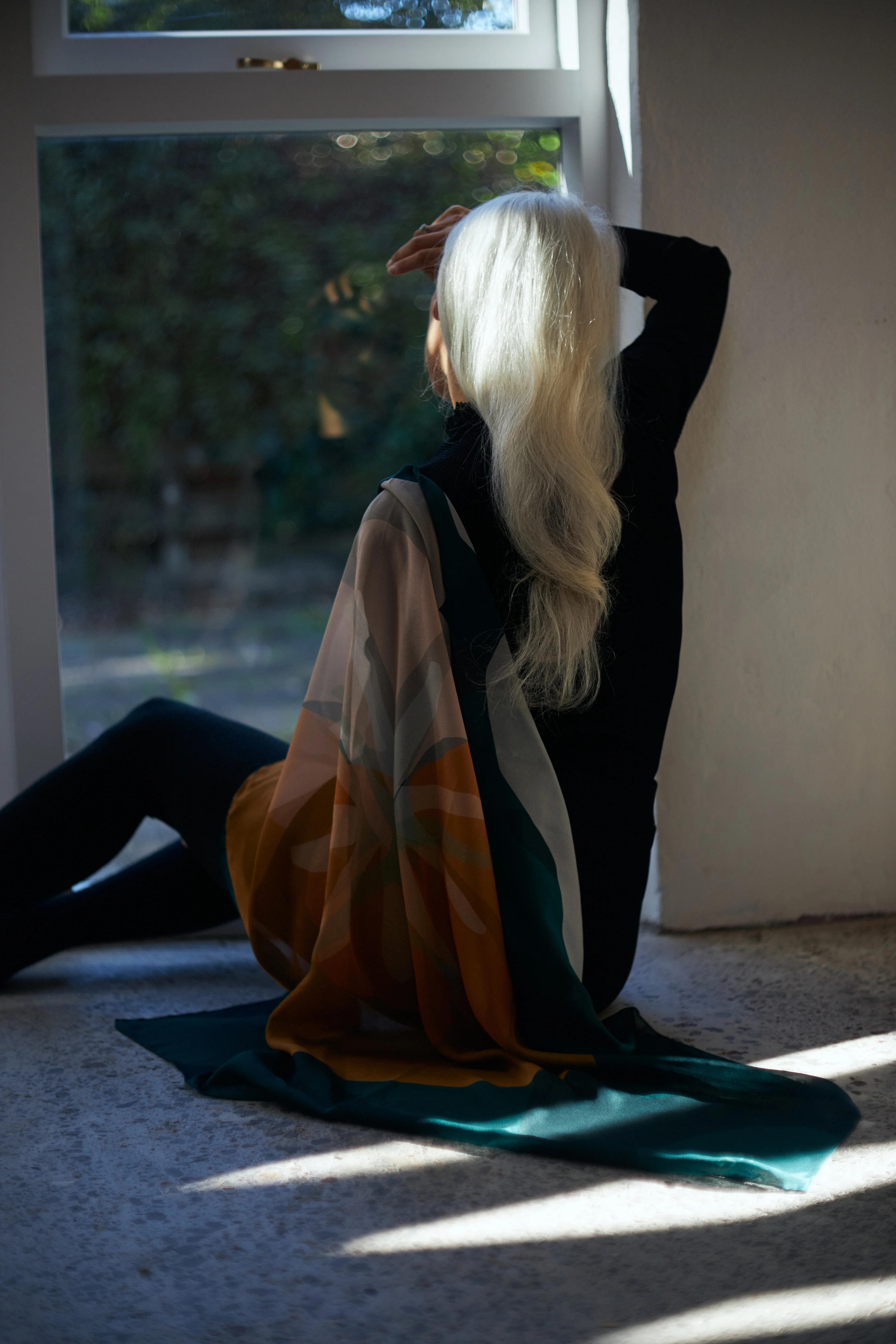 A person with long white hair sits on the floor in front of a large window, draped in a colourful shawl. They hold one arm up toward the window, shielding their eyes from the light, creating a reflective and serene atmosphere.