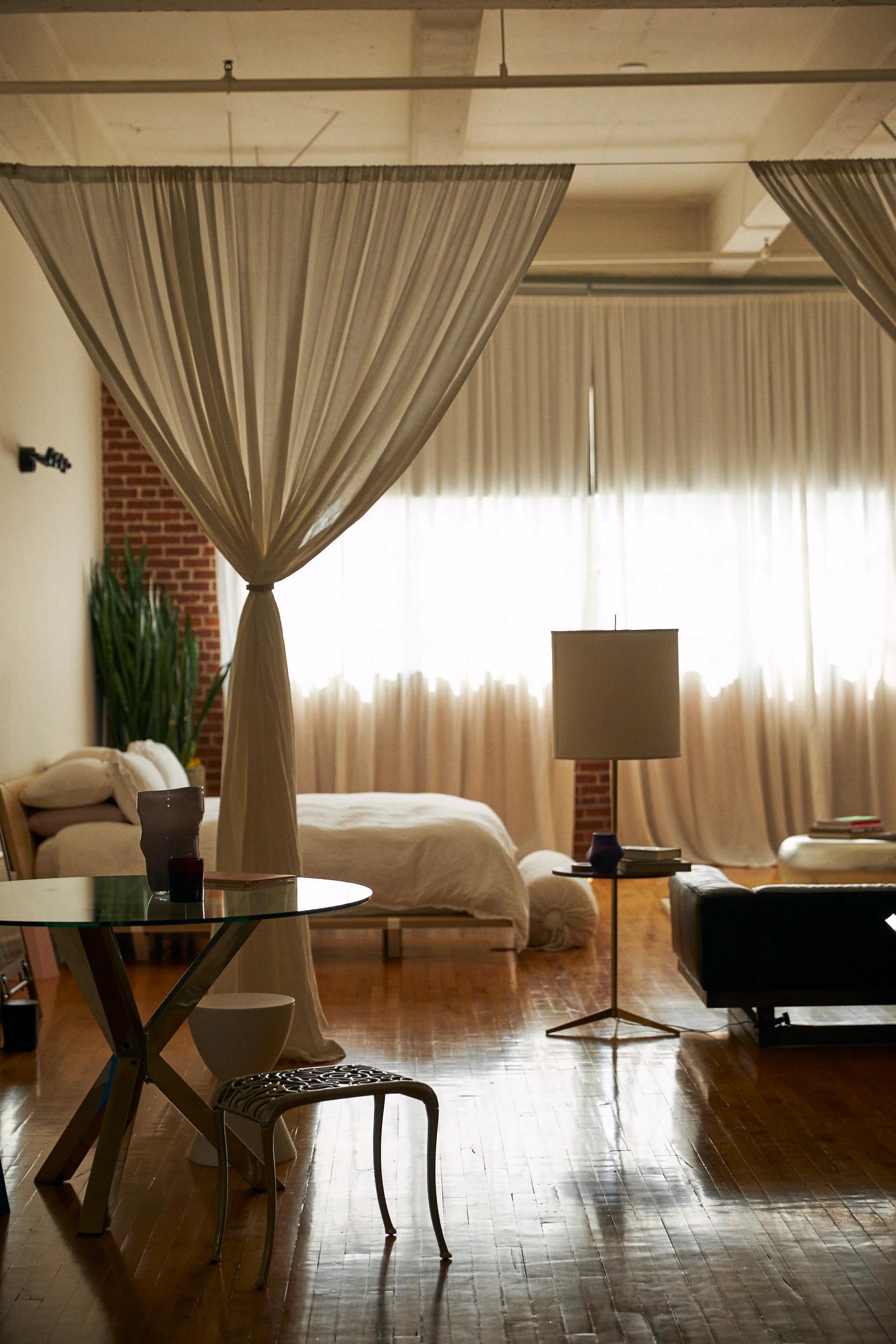 Cozy bedroom with curtains, large bed, and plant. Circular table, chair, and black sofa. Natural light filters through.