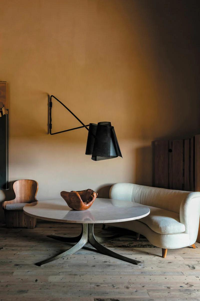 A Provençal-inspired room with a round wooden table, modern sofa, and chair. A black industrial light hangs above, creating a warm ambiance.