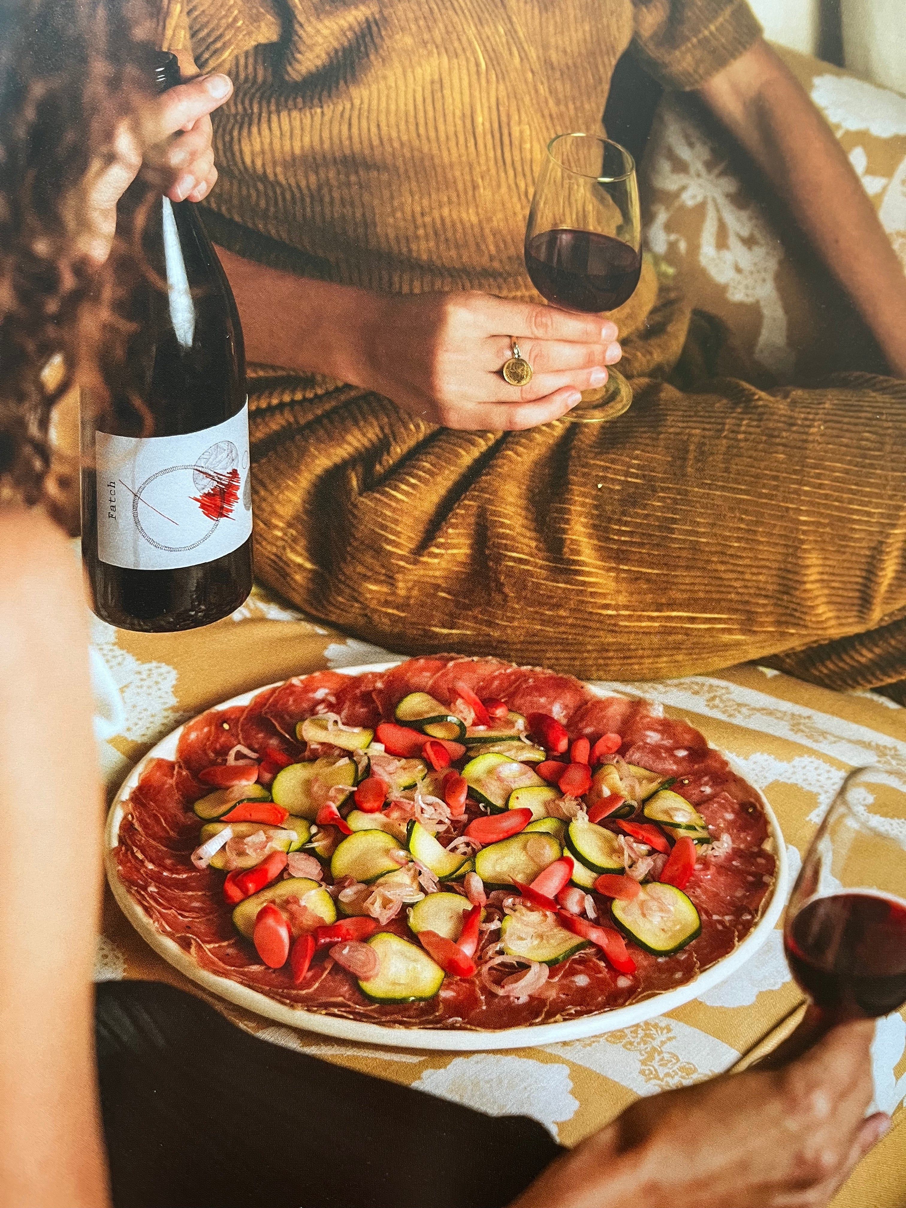 Two people with wine glasses and bottle. Plate of cured meats with cucumbers and radishes. Seated on patterned blanket or sofa.