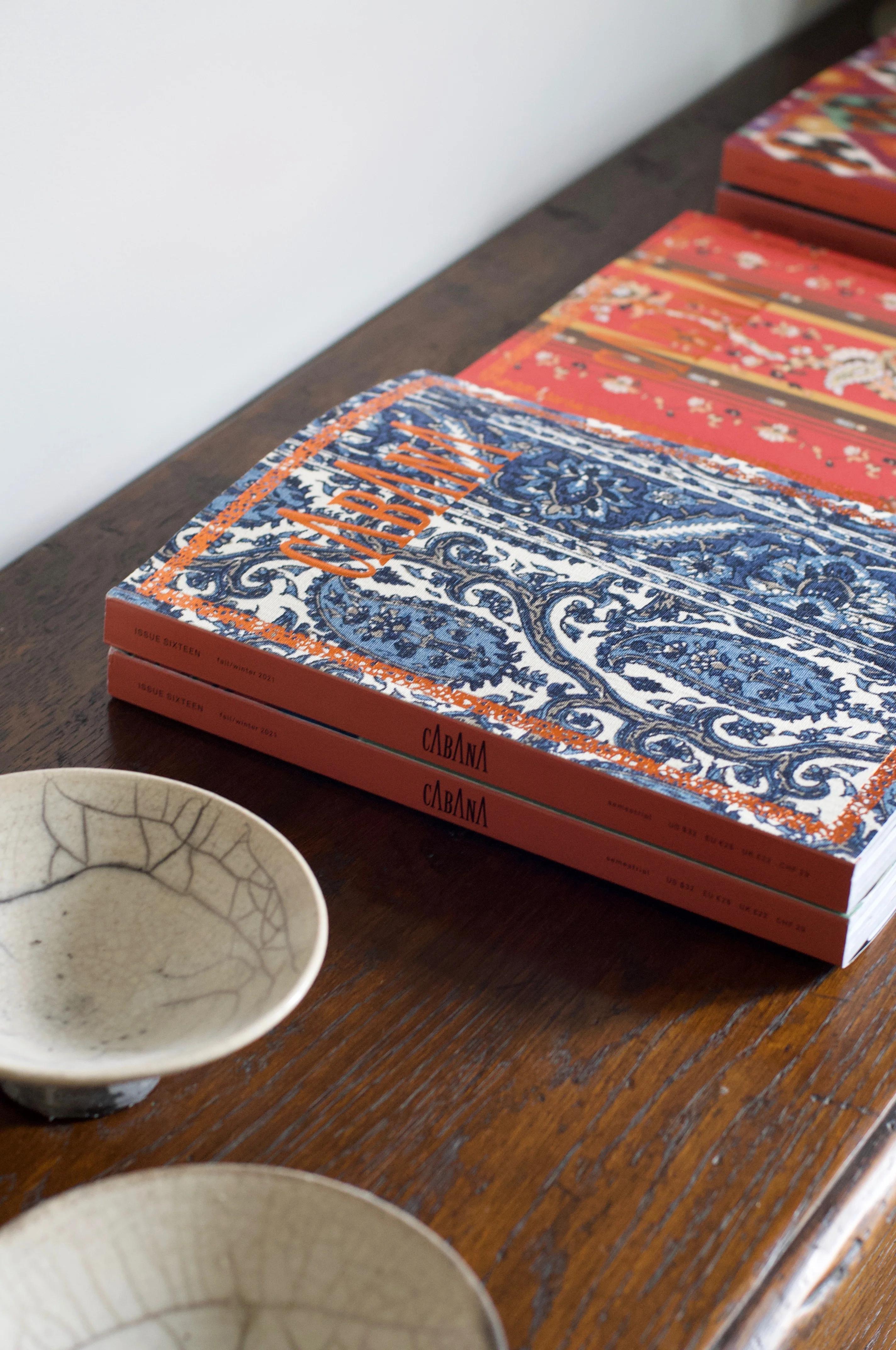 Two CABANA Magazine stacks on wood. Top issues show blue-white and red-orange patterns. Crackle-glazed bowls in foreground.