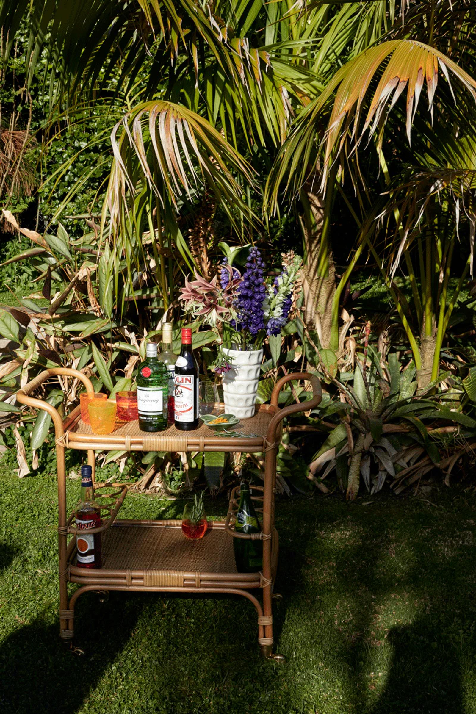 Bamboo bar cart on lawn with bottles, glasses, and purple flowers. Surrounded by tropical plants in sunlit setting.