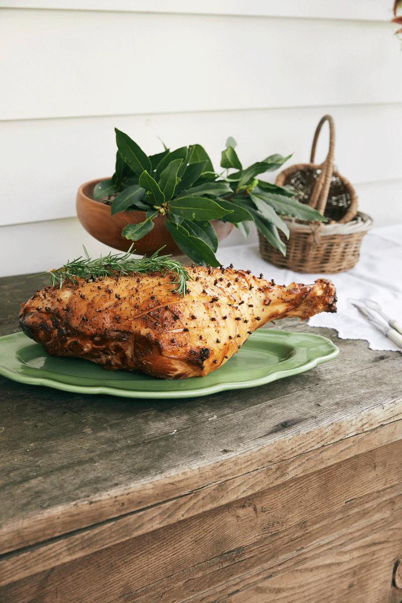 A roasted Christmas Ham garnished with rosemary and cloves on a green platter. A basket and bowl of greens in the background evoke a cozy atmosphere.