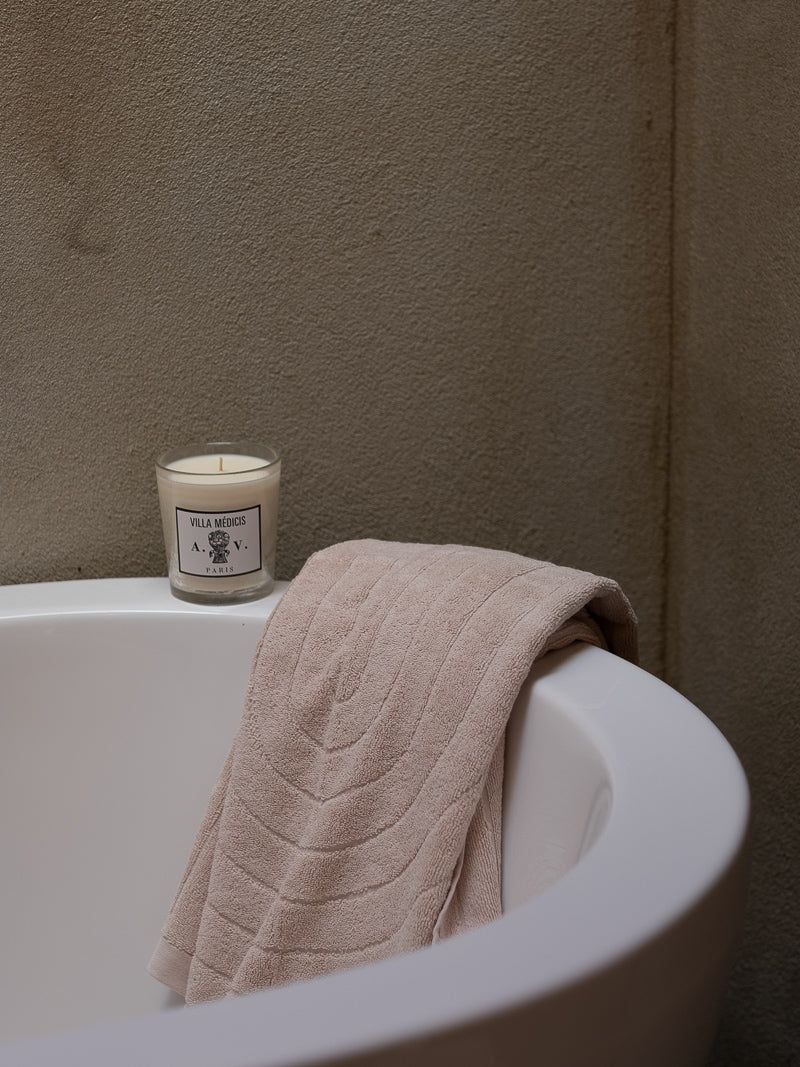 A white bathtub with a beige towel draped over the edge and a "Villa Medici" candle on the edge. A grey textured wall background creates a serene atmosphere.