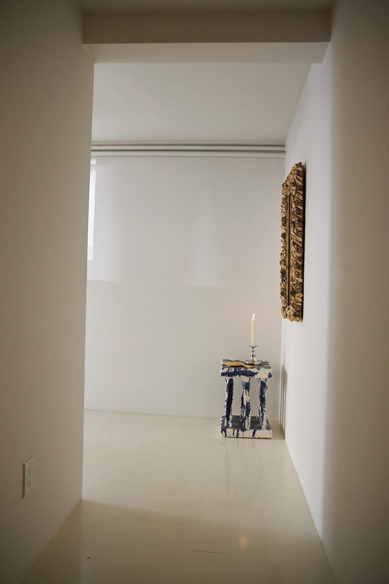 Minimalist room with white walls. Narrow table with blue and white ceramics. Lit candle on table. Textured artwork on wall.