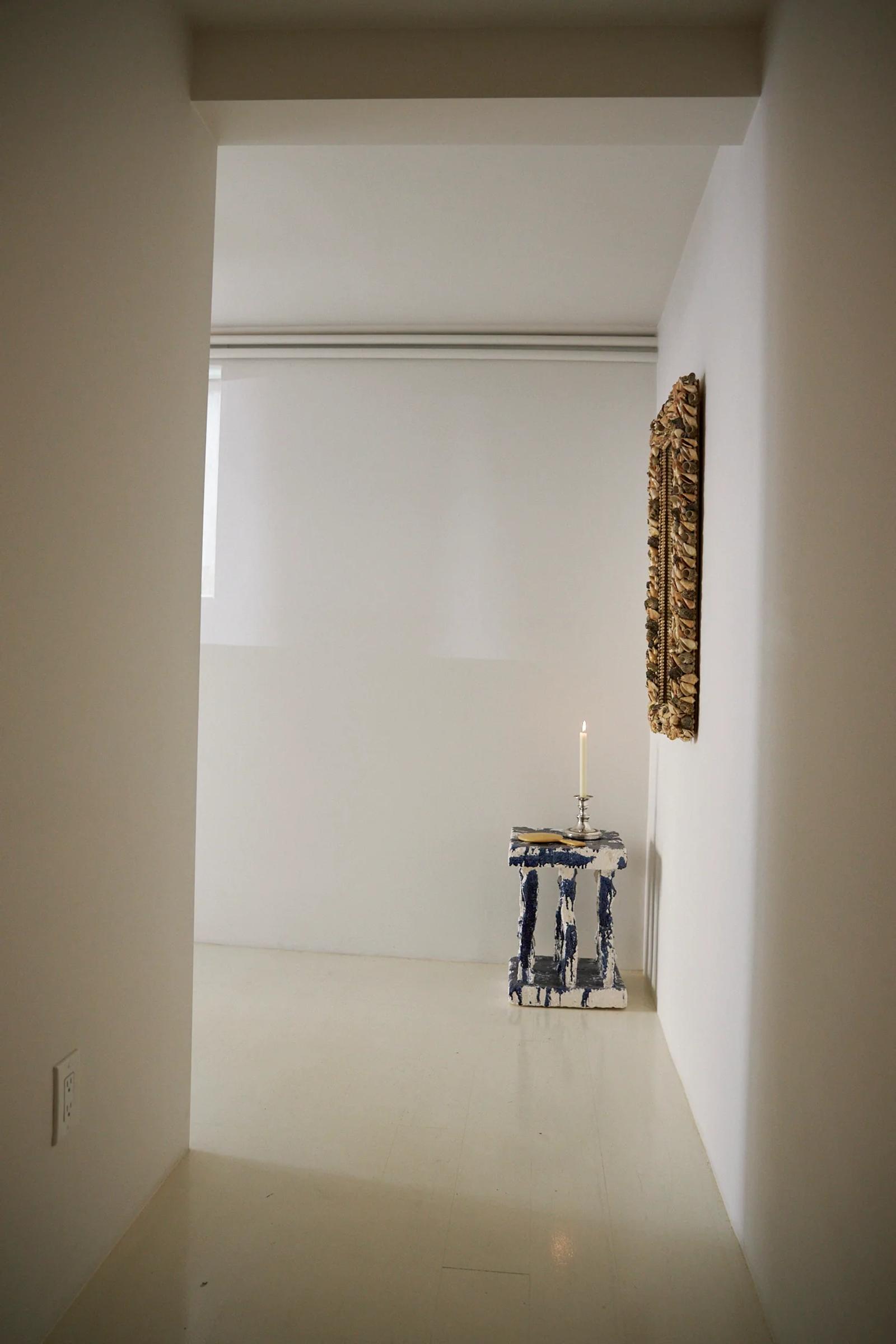 Minimalist room with white walls. Narrow table with blue and white ceramics. Lit candle on table. Textured artwork on wall.
