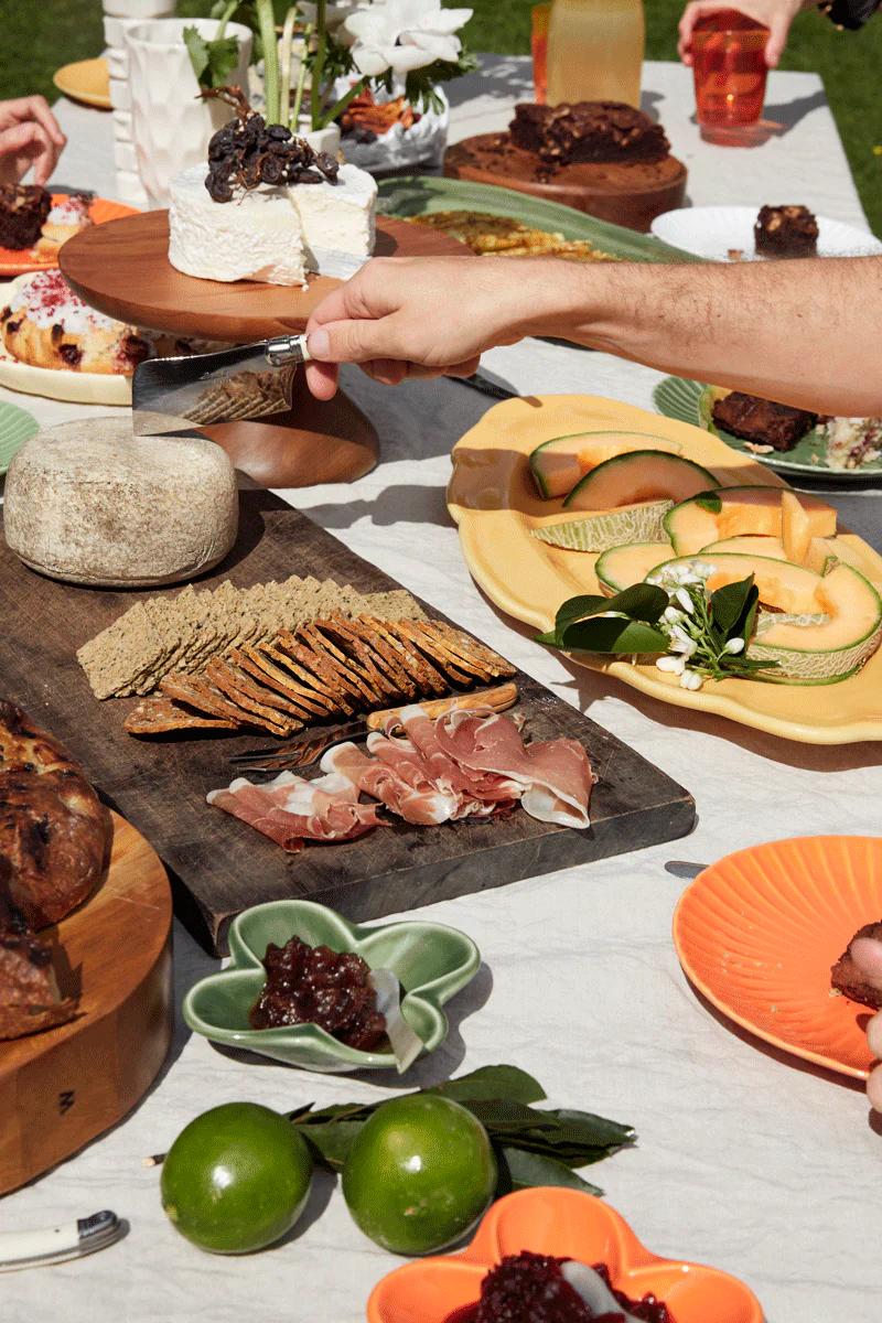 A hand cuts a cheese wheel at an outdoor table set with cheese, crackers, meats, fruit, and drinks, perfect for al fresco dining.