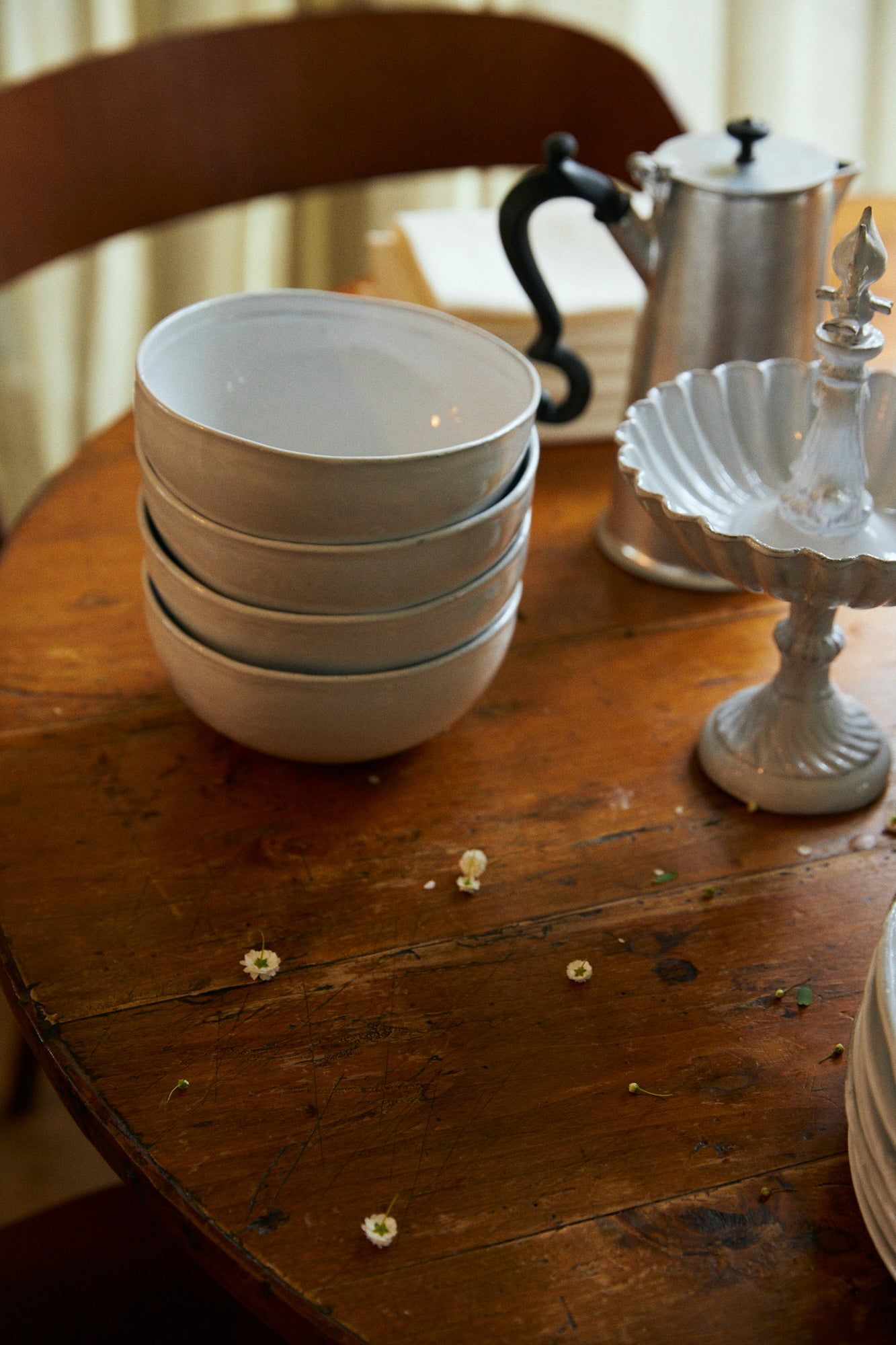 Rustic table with stacked bowls, silver teapot, and white tiered stand. Small white flowers scattered on table.