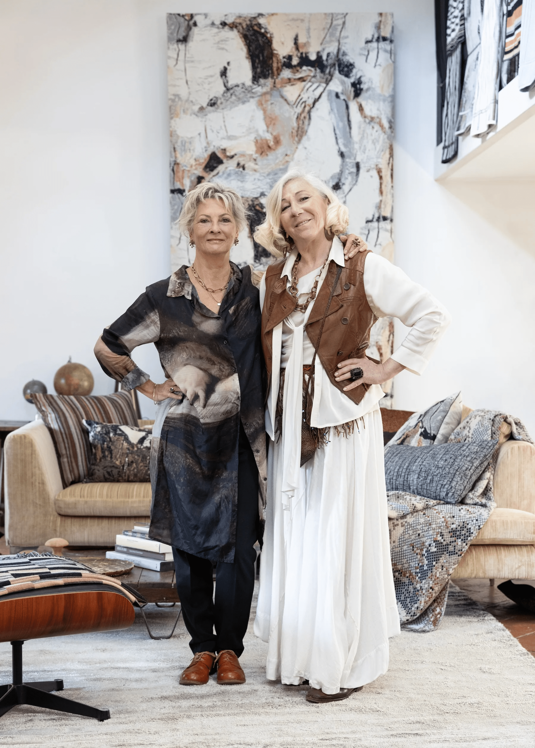 Two women in stylish living room. One in artistic tunic, other in white gown with brown vest. Abstract painting behind.