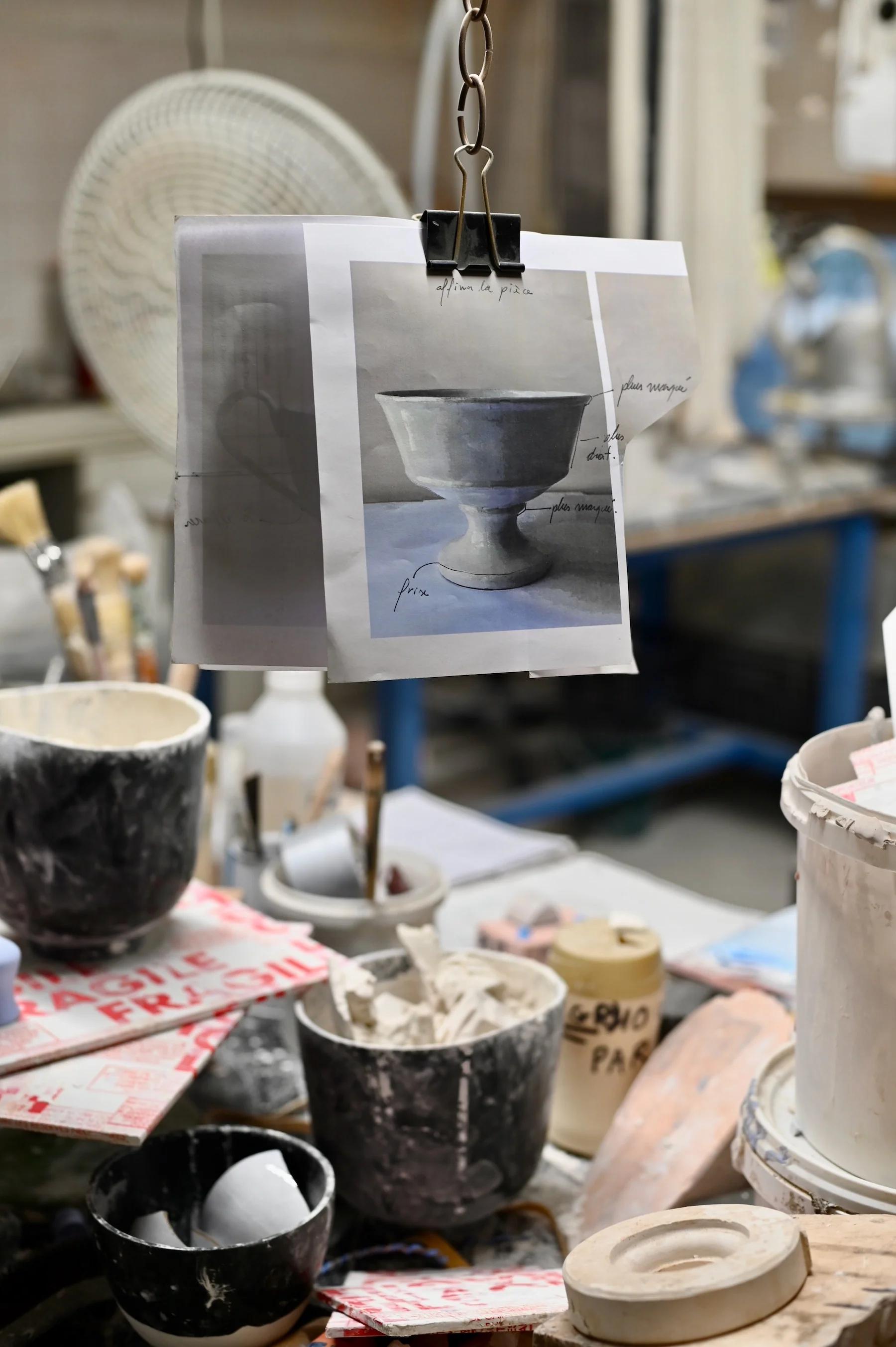 Cluttered pottery studio with various tools, supplies, and ceramic pieces. Design sketch of bowl hanging from clip in center.