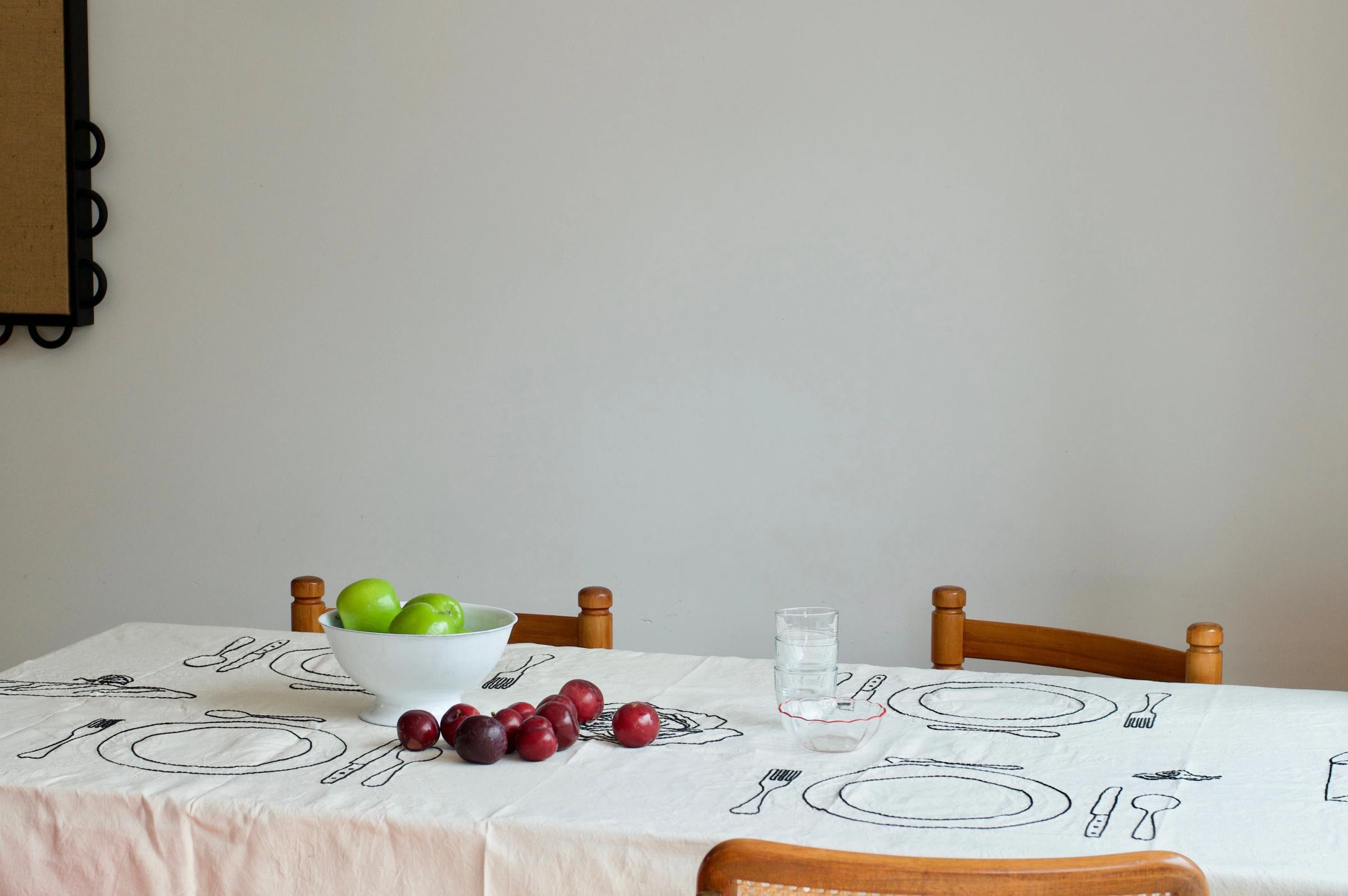 Table with embroidered linens, bowl of green apples, red apples and plums, clear water glass. Four wooden chairs surround.