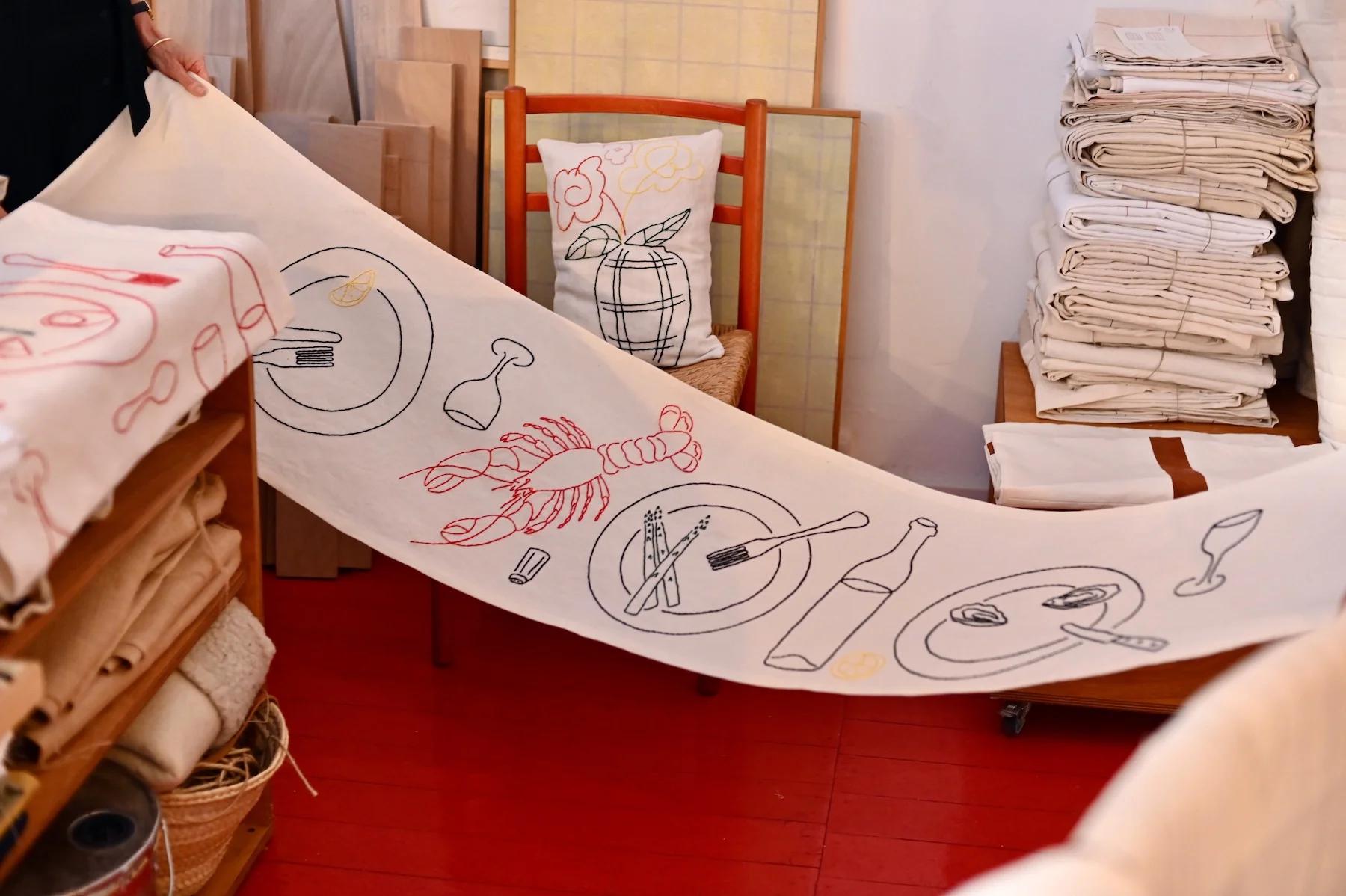 Person unrolls fabric with colourful kitchen item drawings. Room has stacked fabrics, baskets, and wooden furniture on red floor.