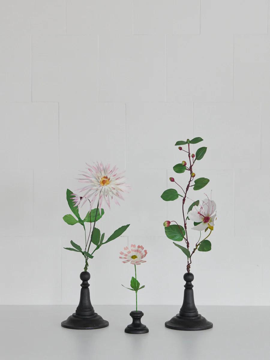 Three flower arrangements in black pedestal vases. Soft pink blooms with green leaves. White tiled background.