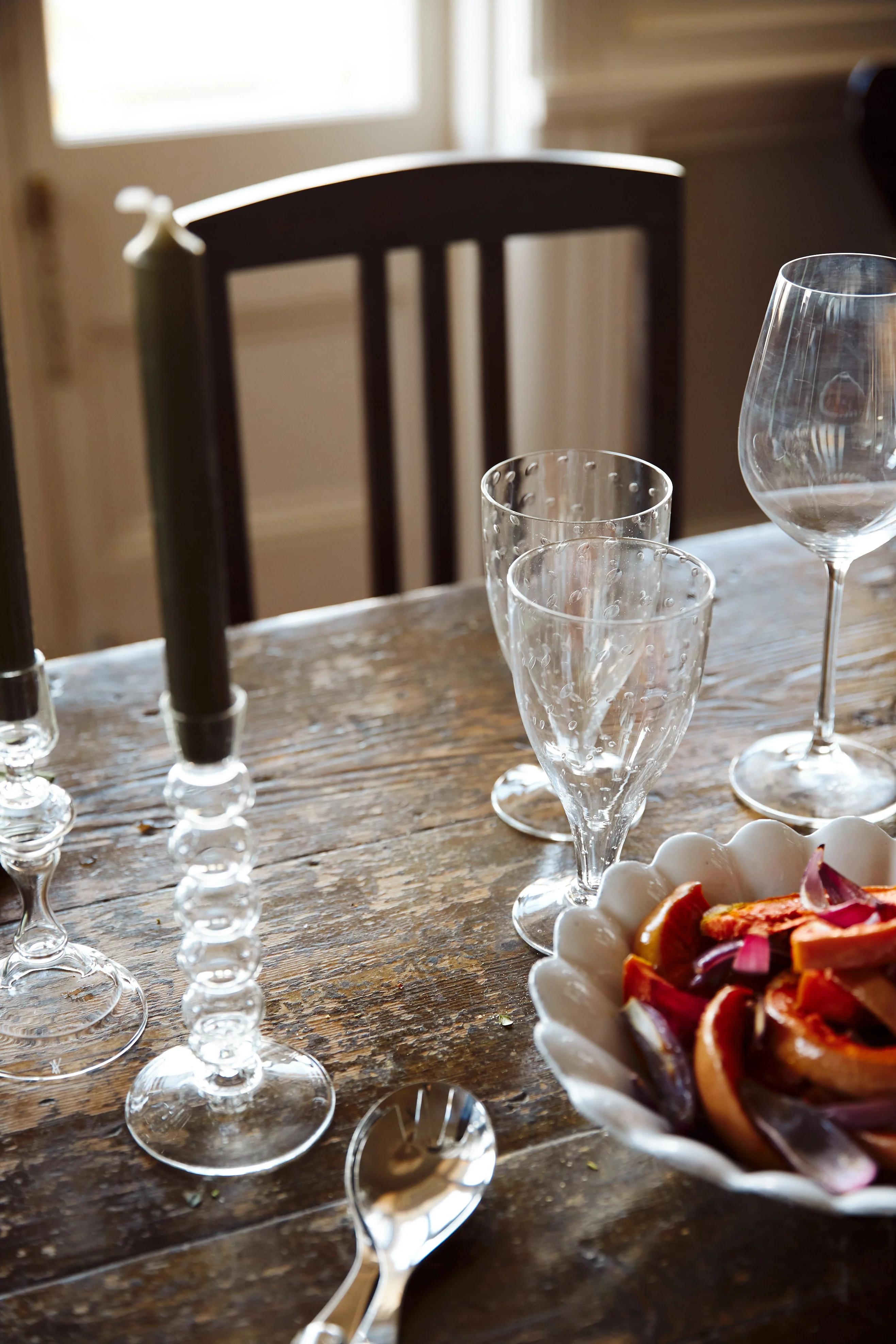 Wooden table with wine glasses, candles, and citrus bowl. Chair and window in background.