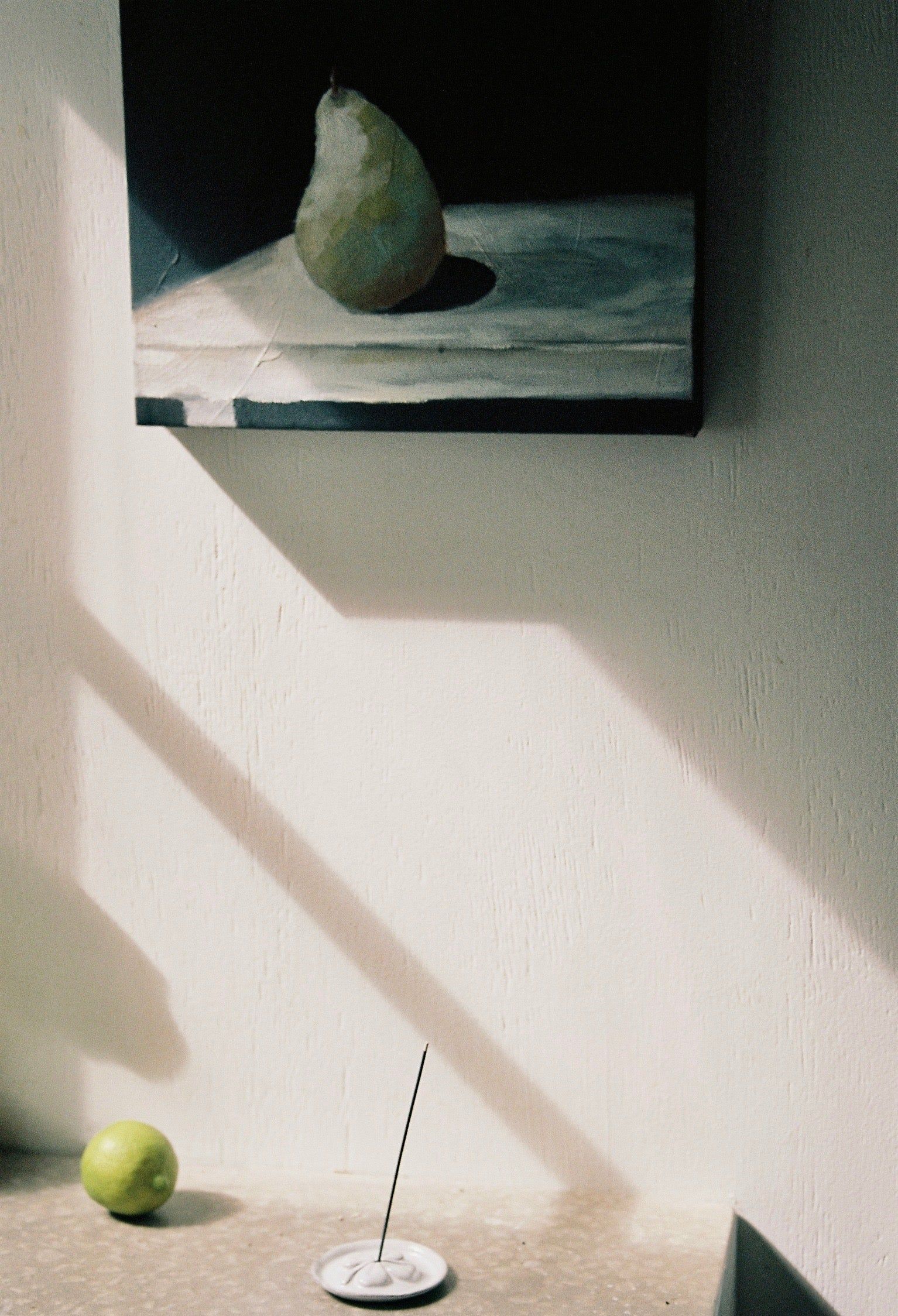 A painting of a pear on a sunlit wall, with an incense stick and a lime on a ledge, creating a peaceful ambiance in a seaside cottage.