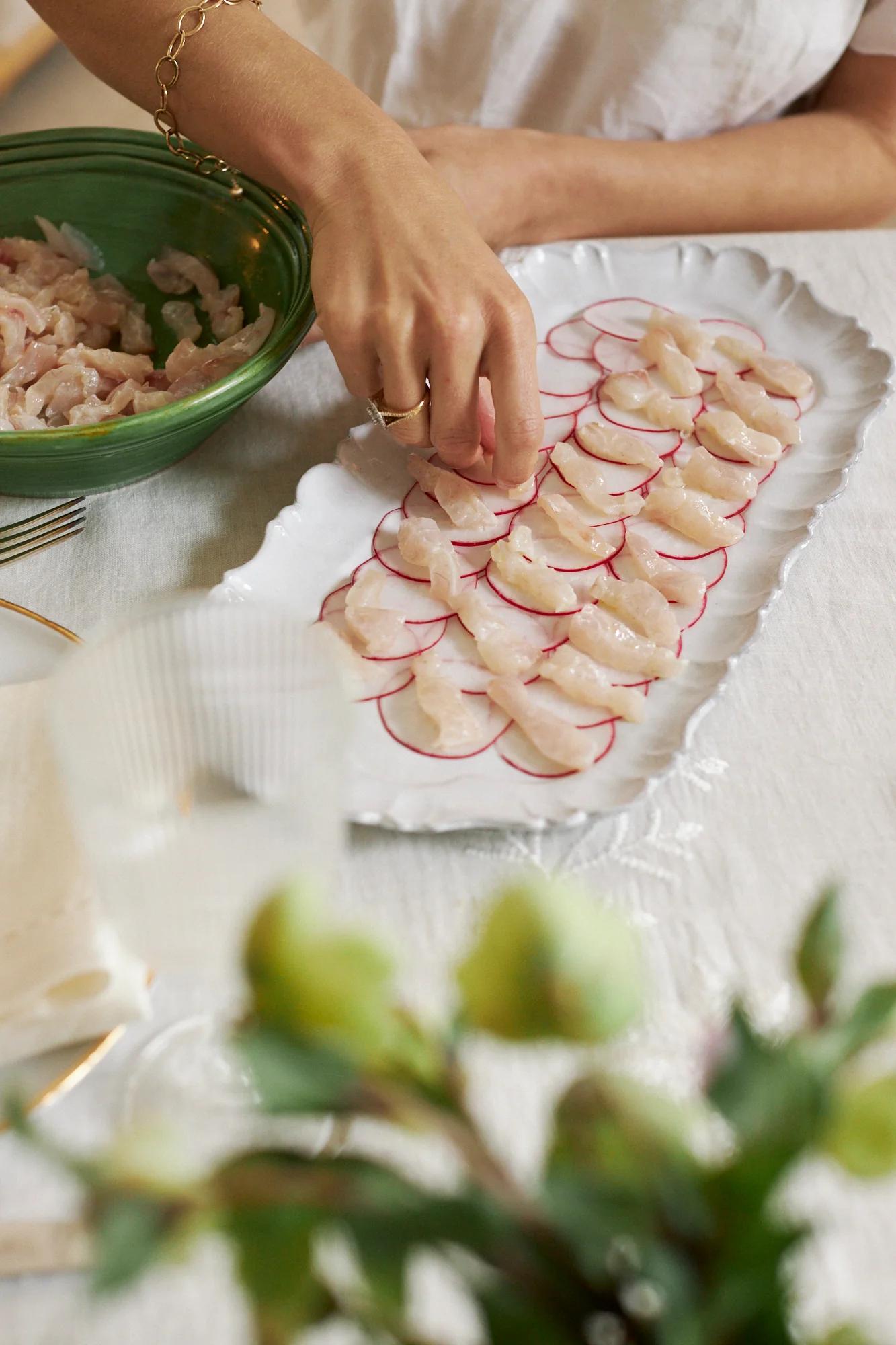 A person plates raw fish and radish on a white platter. A green bowl, glass, and blurred flowers complete the French-inspired table setting.