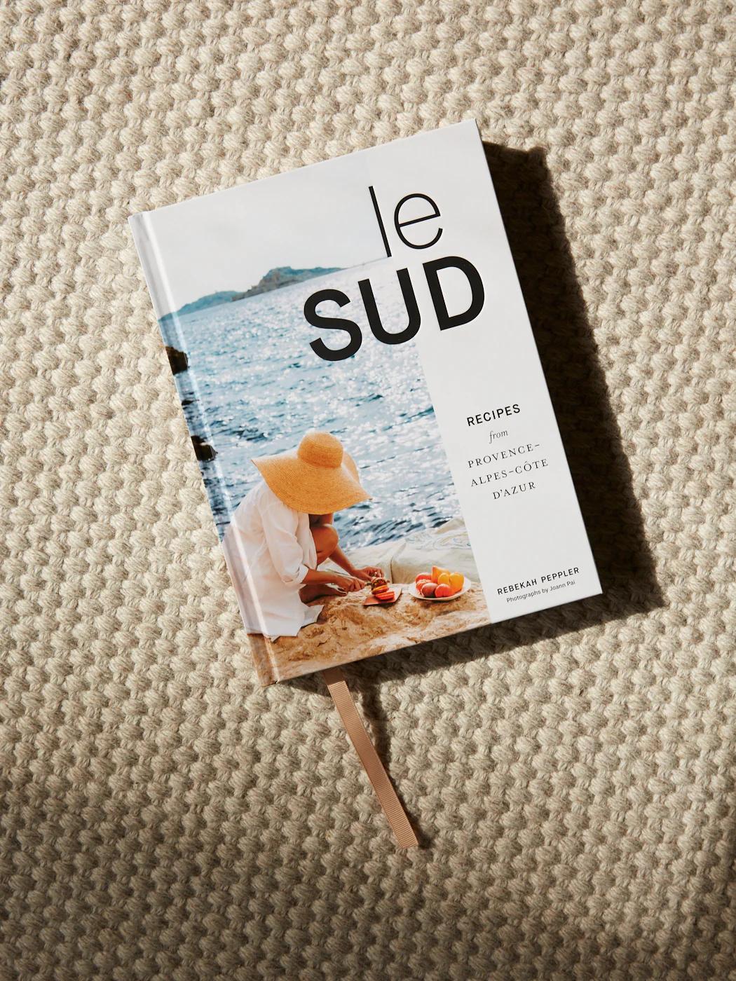 "Le Sud" cookbook on beige surface. Cover shows person in hat by sea arranging produce on beach.