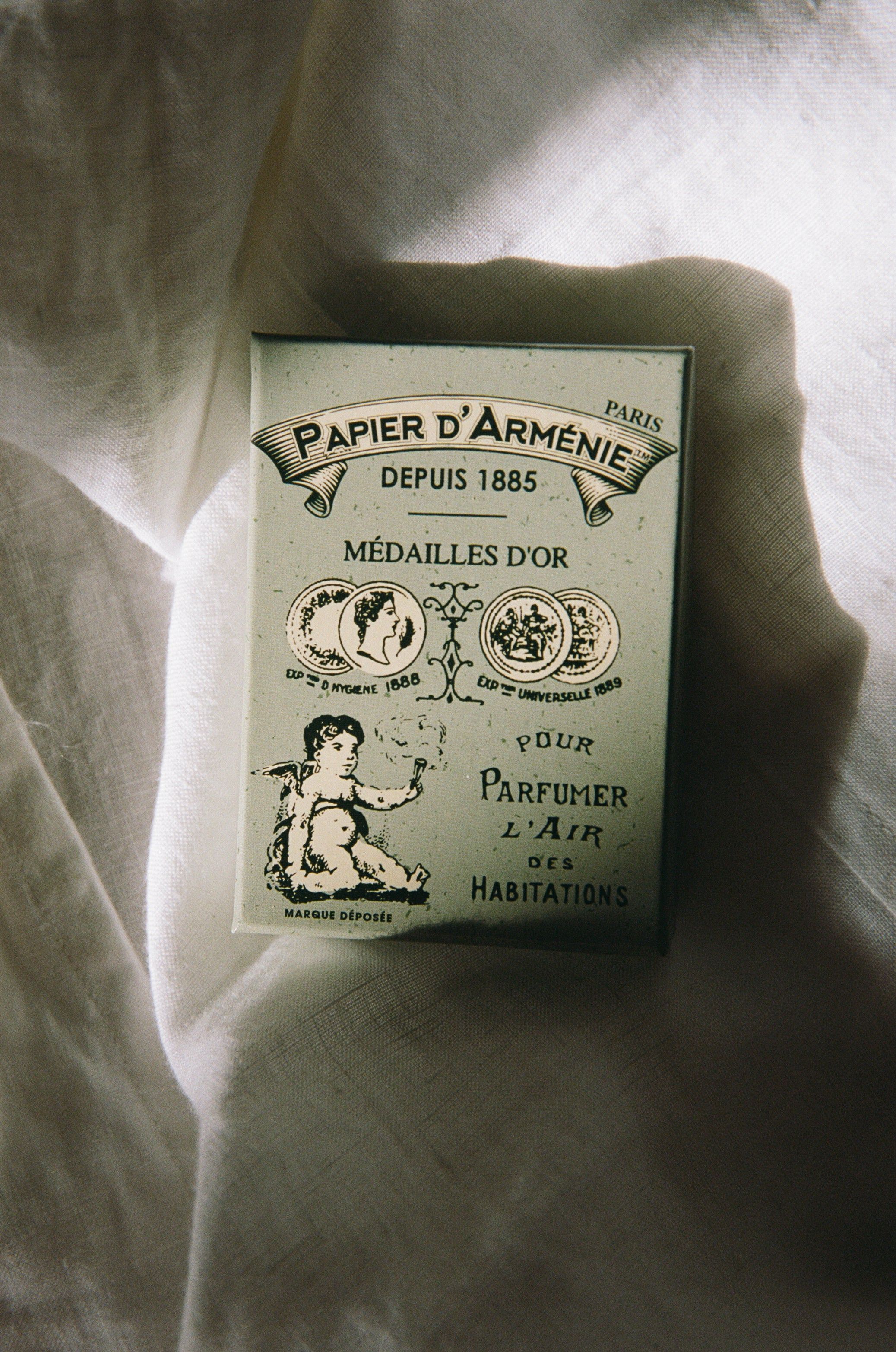 A vintage Papier d’Arménie Paris box with ornate designs and French text rests on crumpled white fabric. The box, featuring gold medals, has enhanced homes’ scents since 1885.