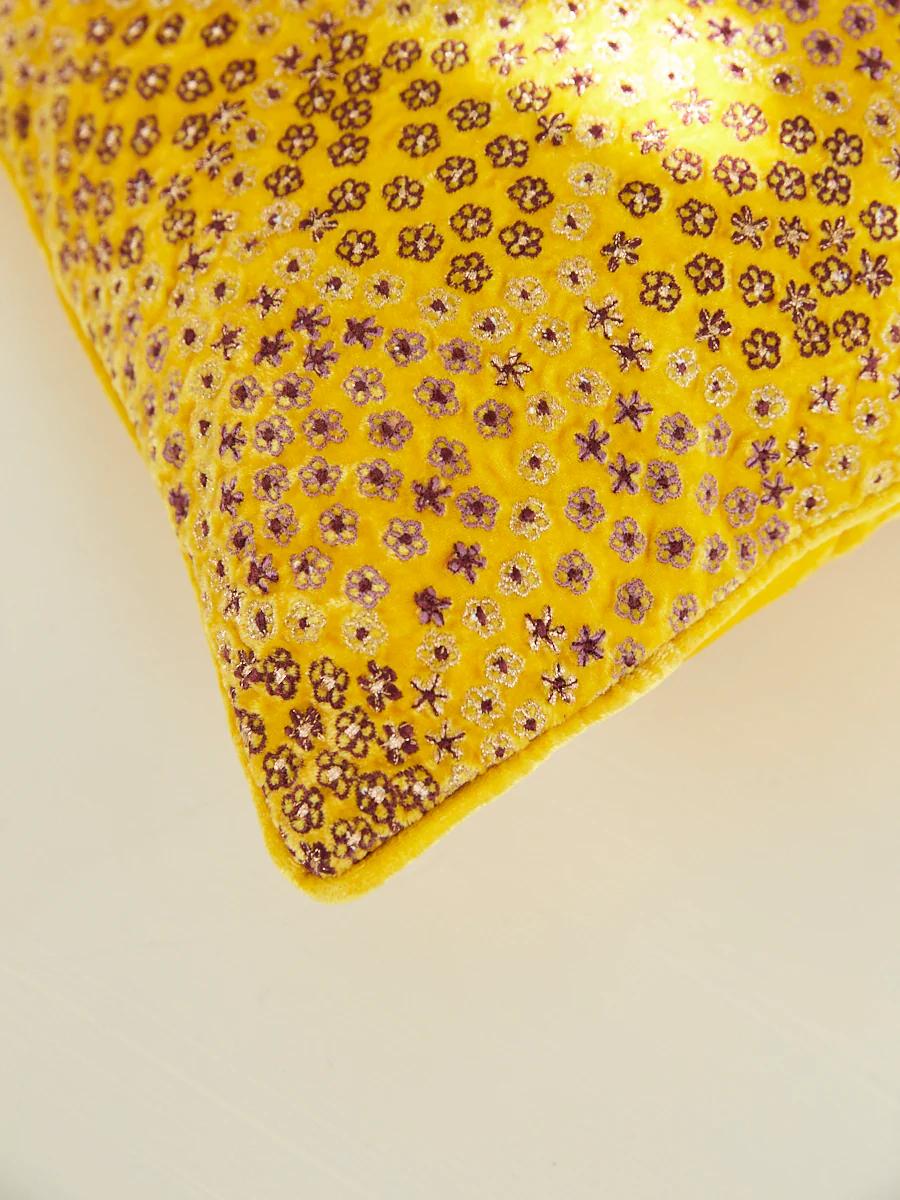 A corner of a bright yellow pillow with intricate floral embroidery is shown. Small purple and gold flowers stand out against the soft, vibrant background.