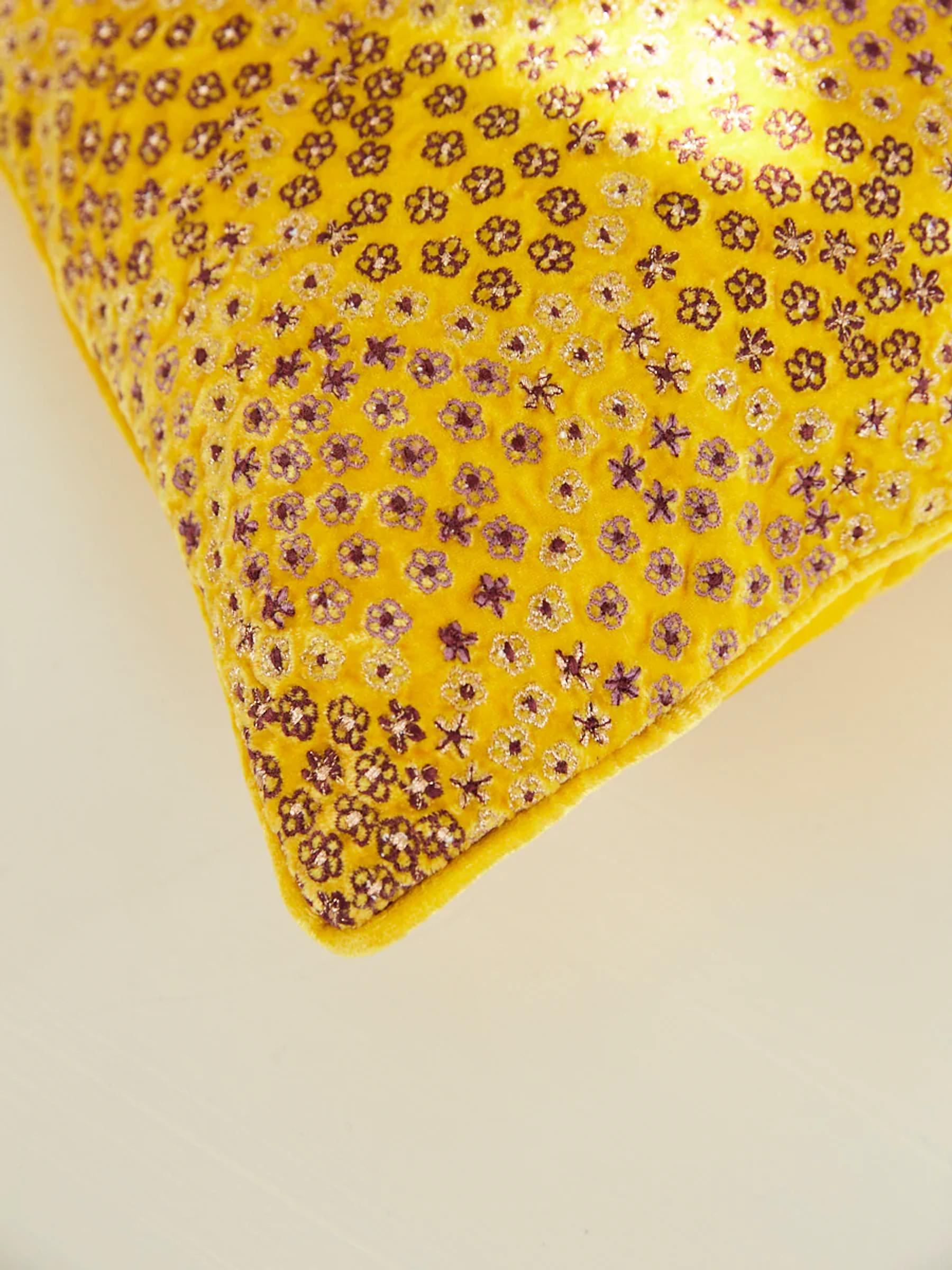 A corner of a bright yellow pillow with intricate floral embroidery is shown. Small purple and gold flowers stand out against the soft, vibrant background.