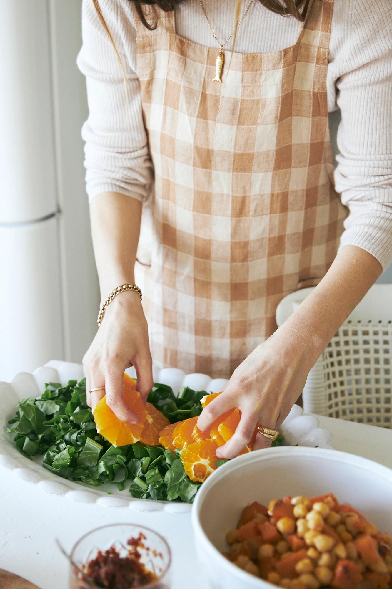 Person in checkered apron making salad with greens, orange slices. Bowls of ingredients nearby.