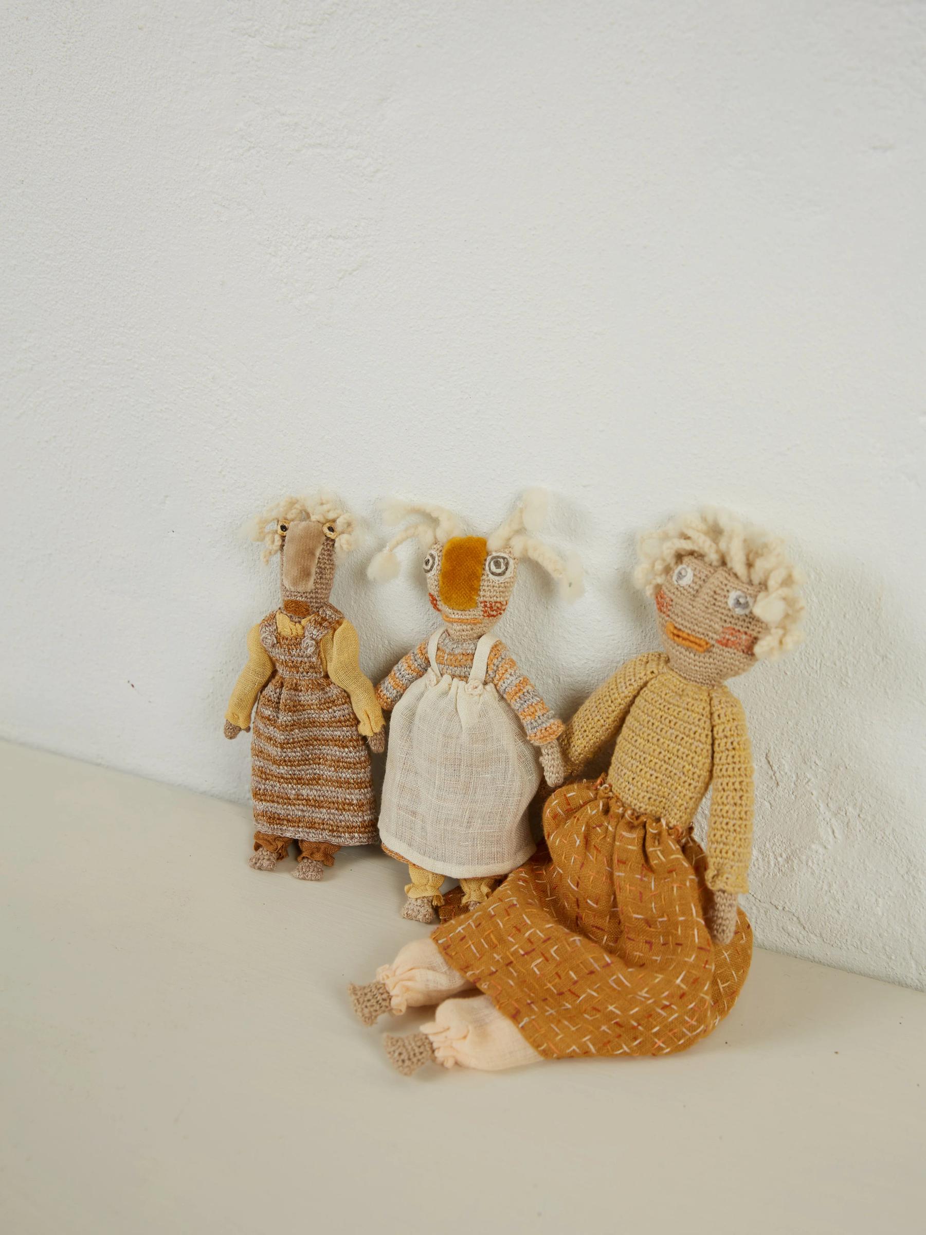 Three handmade dolls against textured white wall. Sophie Digard-style crochet with textured hair and beige clothing. One seated, two propped up, exuding rustic charm.
