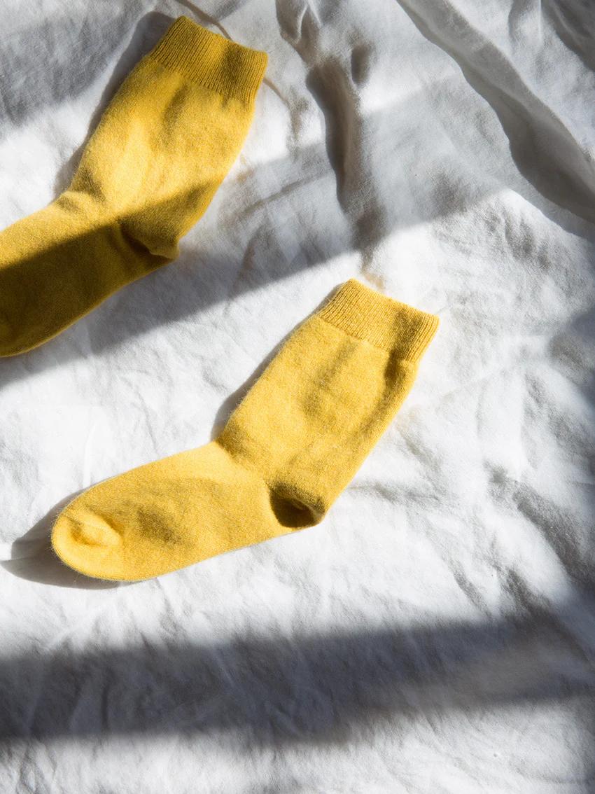 A pair of yellow woollen crew socks on crumpled white fabric. Sunlight casts soft shadows, highlighting the wide ribbing at the top.