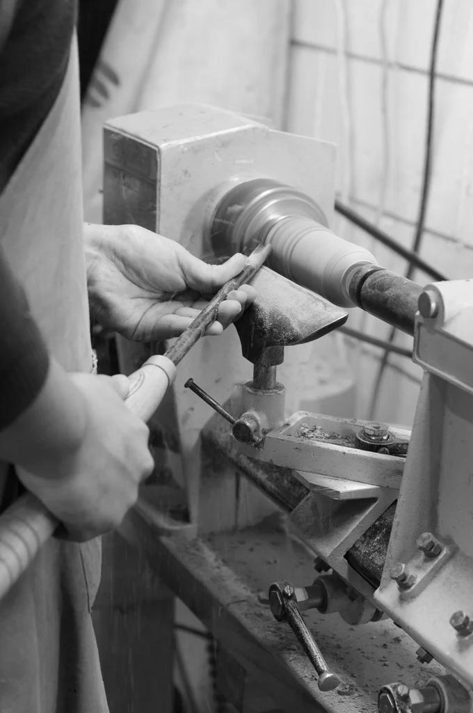 NZ craftsperson shapes wood on lathe, holding chisel. Wearing apron, they work at bench surrounded by tools and wood shavings.