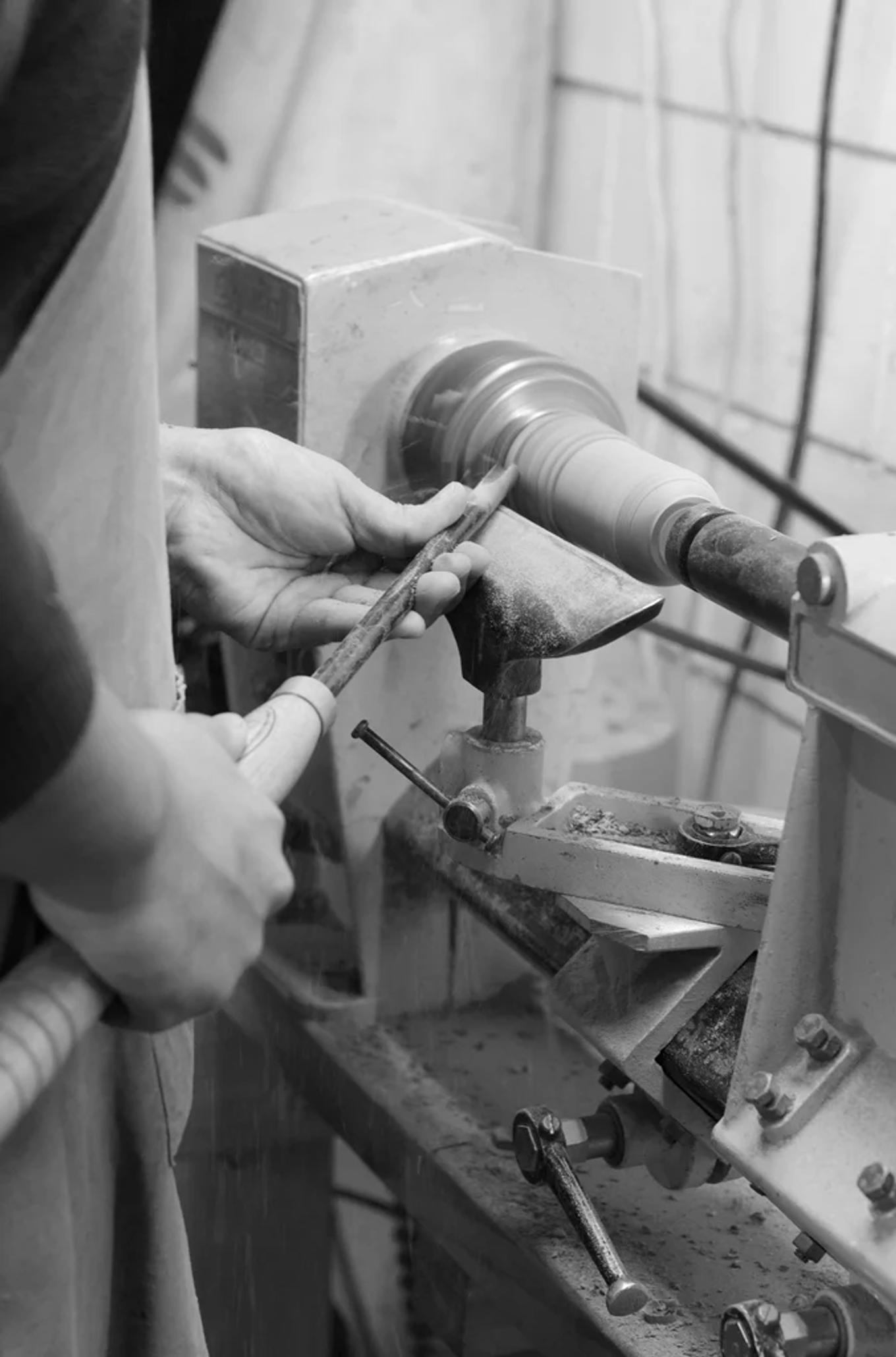 NZ craftsperson shapes wood on lathe, holding chisel. Wearing apron, they work at bench surrounded by tools and wood shavings.