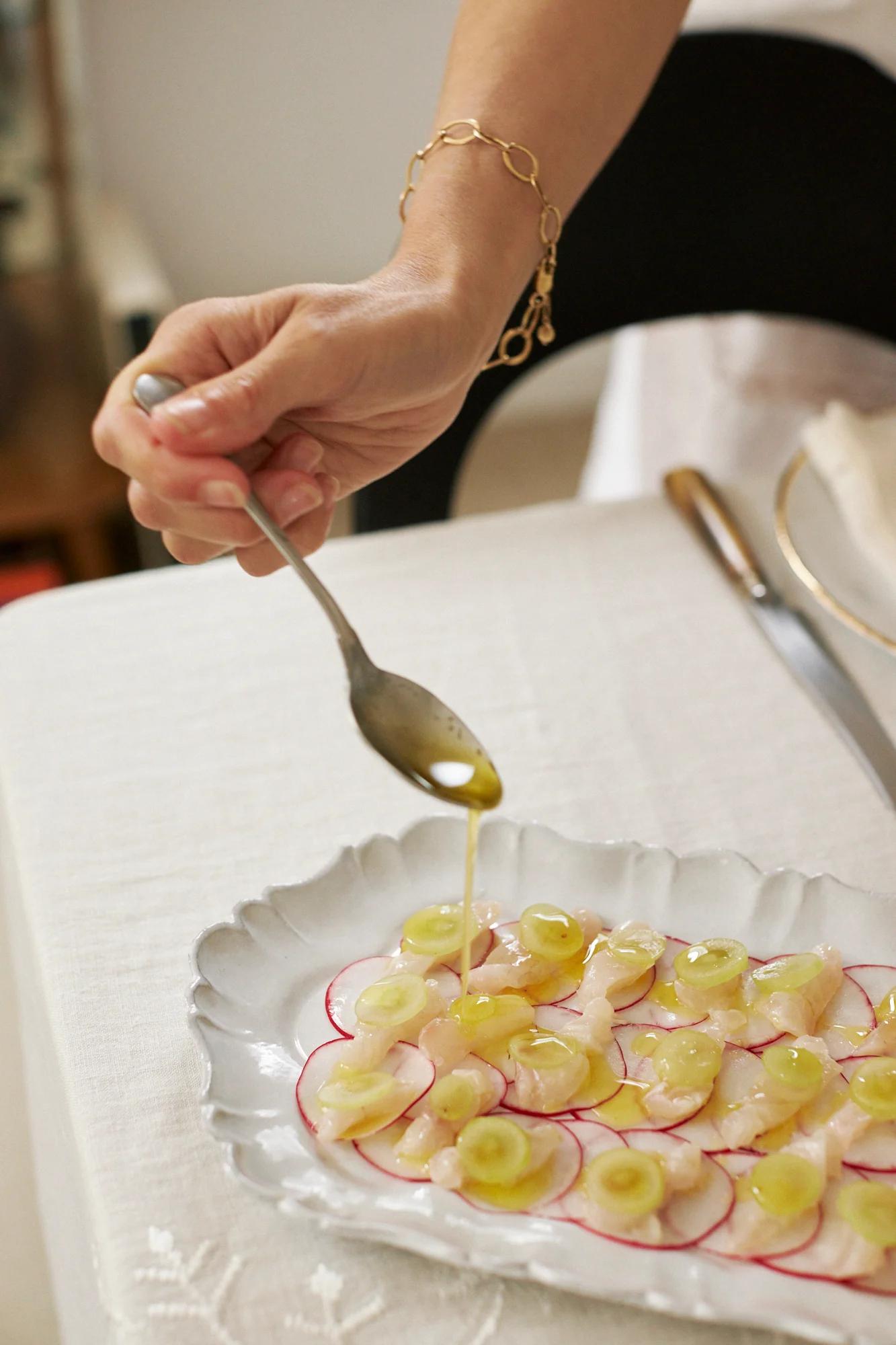 Someone drizzles dressing on sliced radishes and fish. A gold bracelet adorns their wrist. The Marseille-style dish sits with cutlery on a white tablecloth.