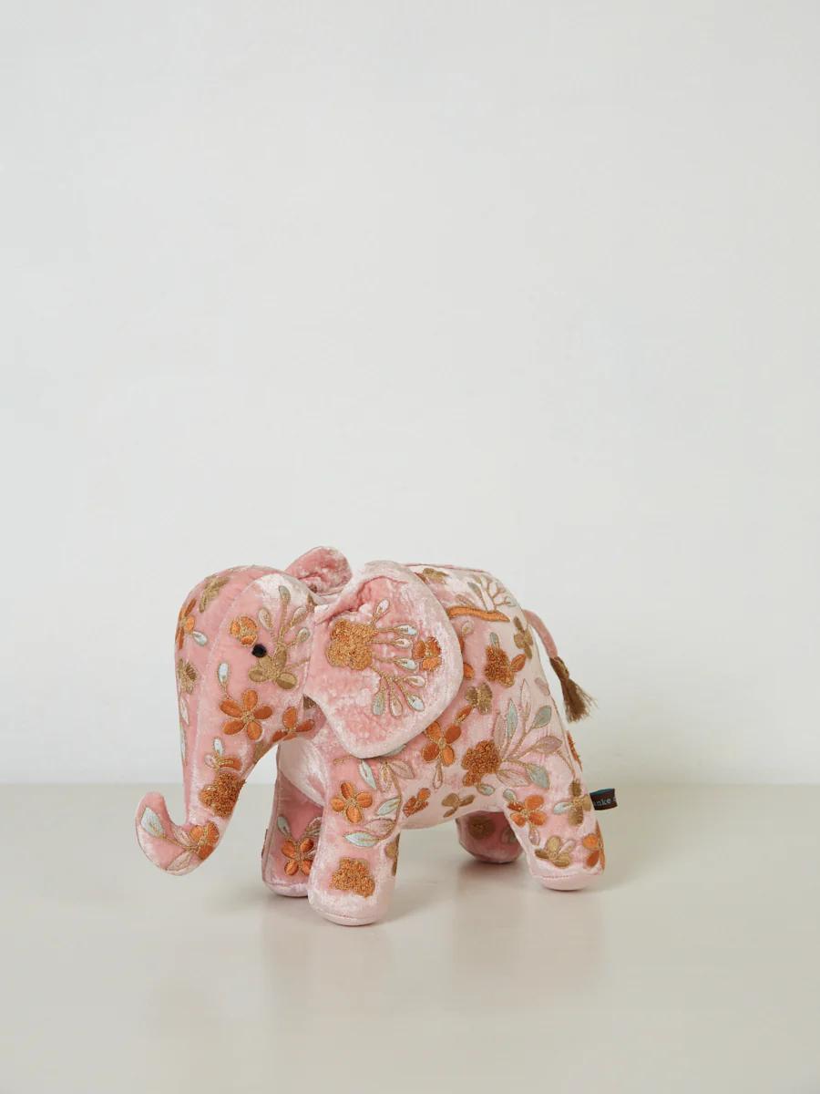 A small pink plush elephant stands on a white surface. Its body is adorned with colourful floral embroidery, showcasing intricate craftsmanship.