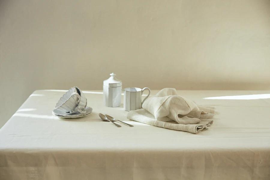 White tablecloth with stacked dishes, Libeco napkin, silver teapot, mug, and spoons. Soft shadows from natural light.