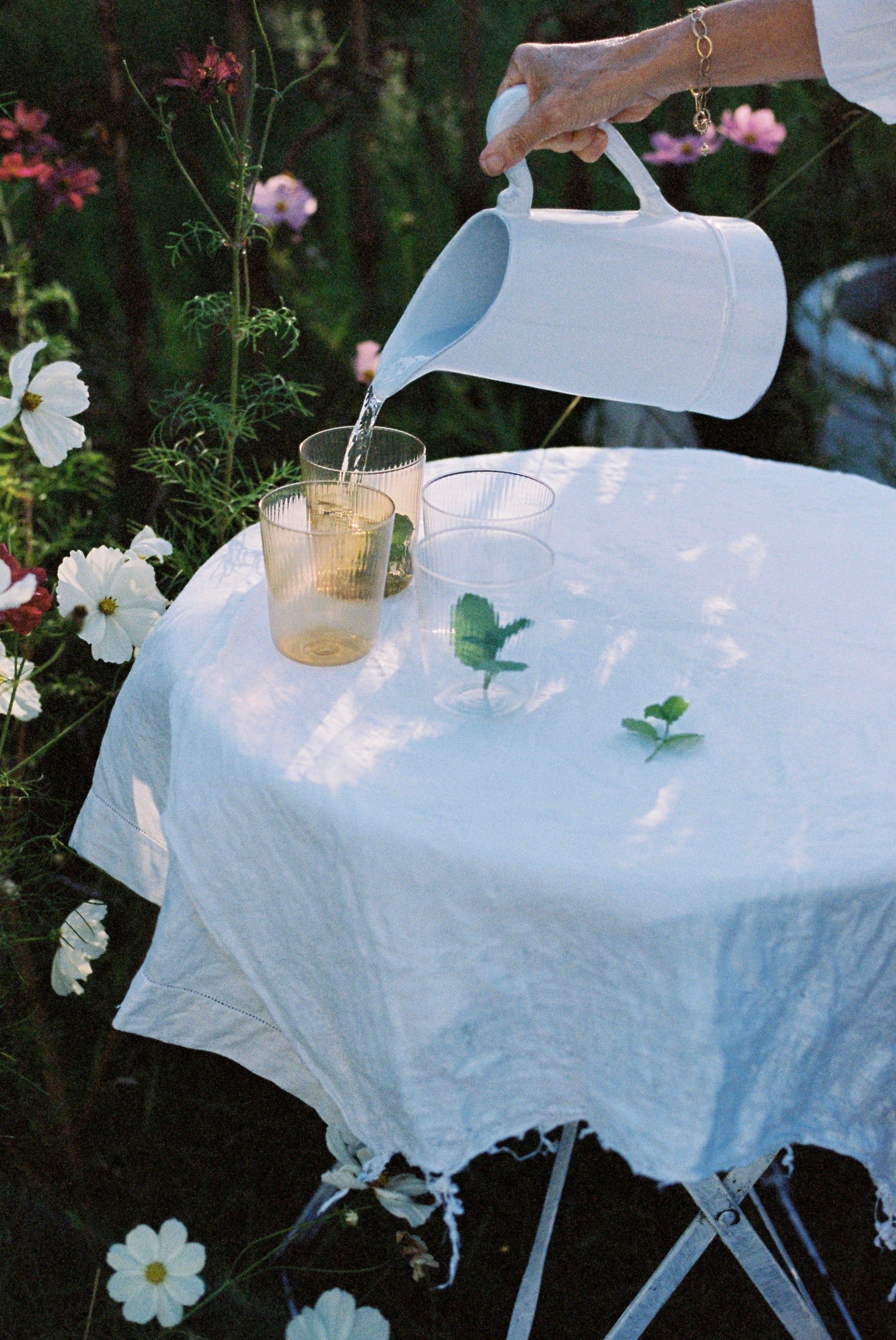 Hand with bracelet pours water from white pitcher into glasses. Table set with white cloth, surrounded by flowers and green frittata, embodying spring in the garden.