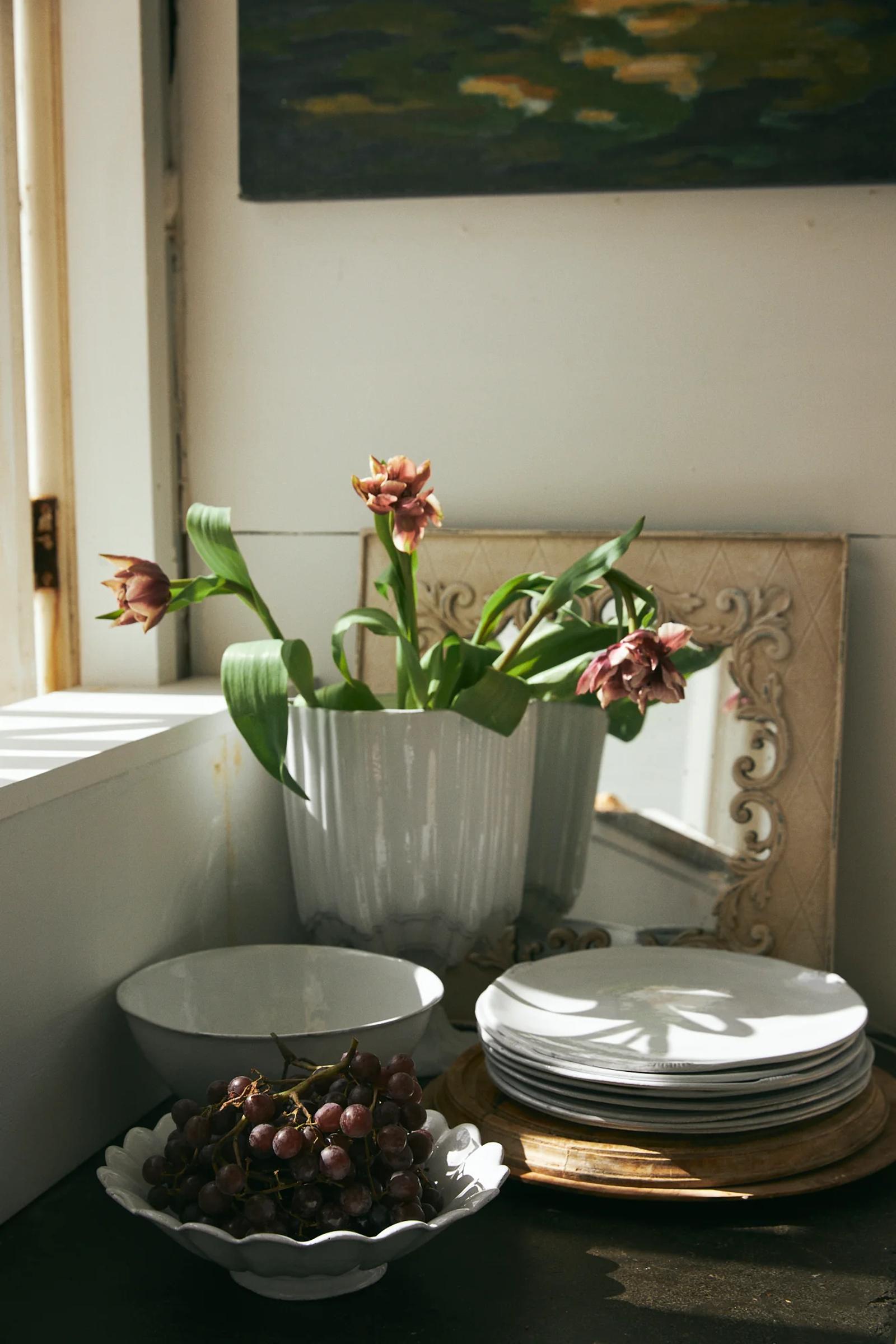 Vase with drooping flowers reflected in mirror. French tableware stacked nearby. Bowl of grapes in foreground. Sunlit window visible.