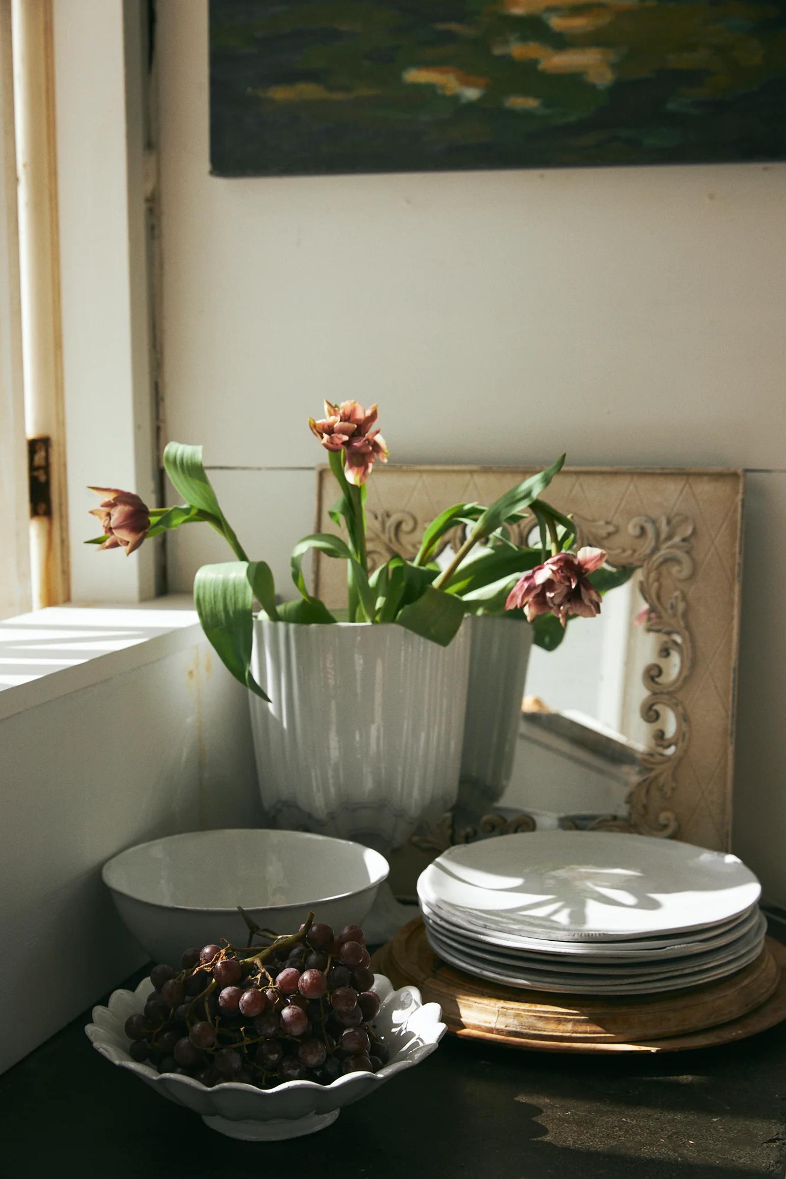 Vase with drooping flowers reflected in mirror. French tableware stacked nearby. Bowl of grapes in foreground. Sunlit window visible.